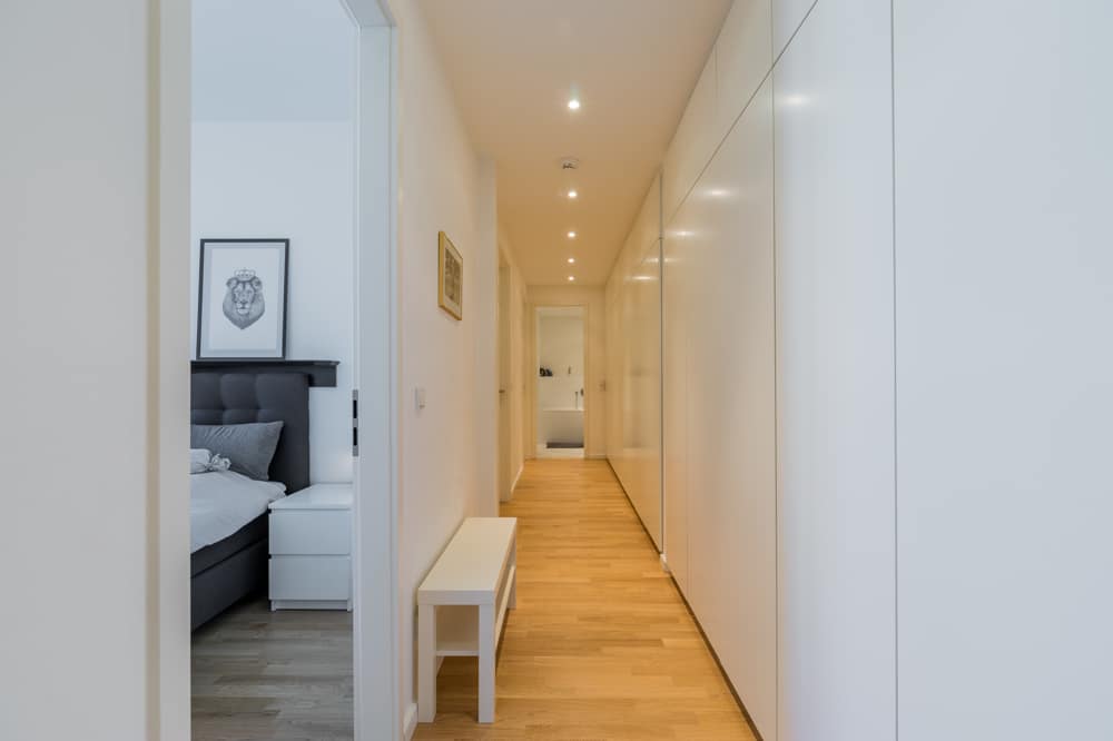 Blick in modernen Apartmentflur mit hellen Einbauschränken, Parkettboden, Deckenspots und Sitzbank neben Schlafzimmer