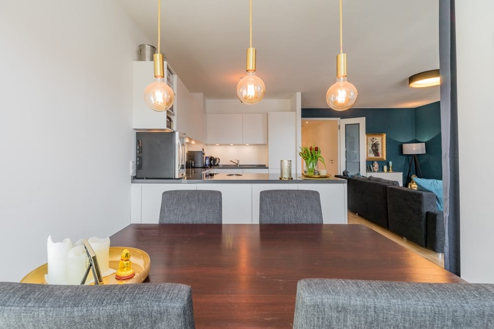 Einblick in offene Wohnküche eines modernen Apartments mit Esstisch, goldenen Pendelleuchten und dunkelgrüner Akzentwand