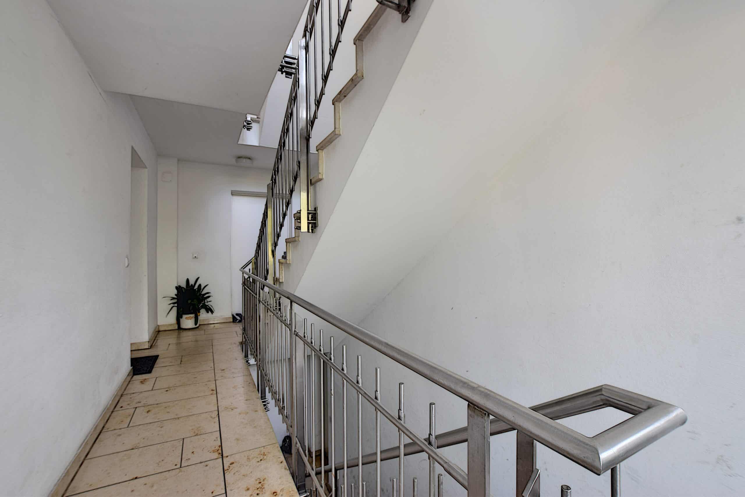 Binnenkant van een woonhuis trappenhuis met roestvrij stalen leuning, tegelvloer, witte muren en een plant