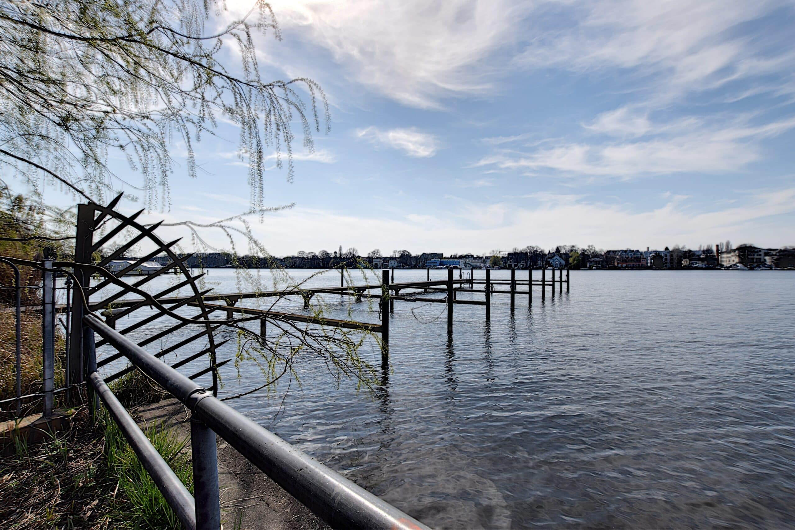 Uitzicht op een meer oever met een oude houten steiger, een gebogen metalen poort en een rij huizen aan de overkant van het water.