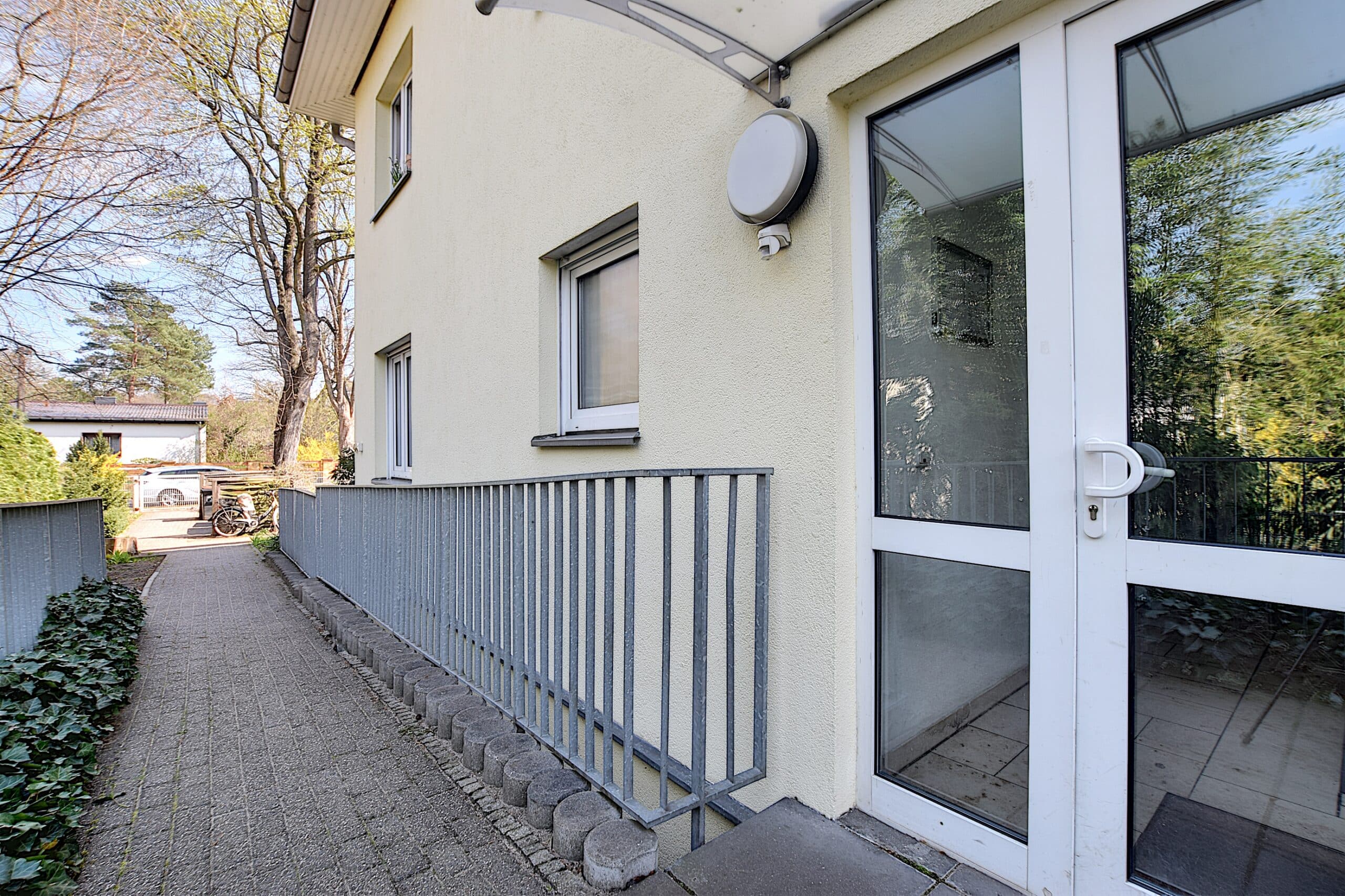 Inzicht op een toegankelijke huisentree met glazen deur en een hellingbaan met metalen leuning tegen een gele pleistergevel.