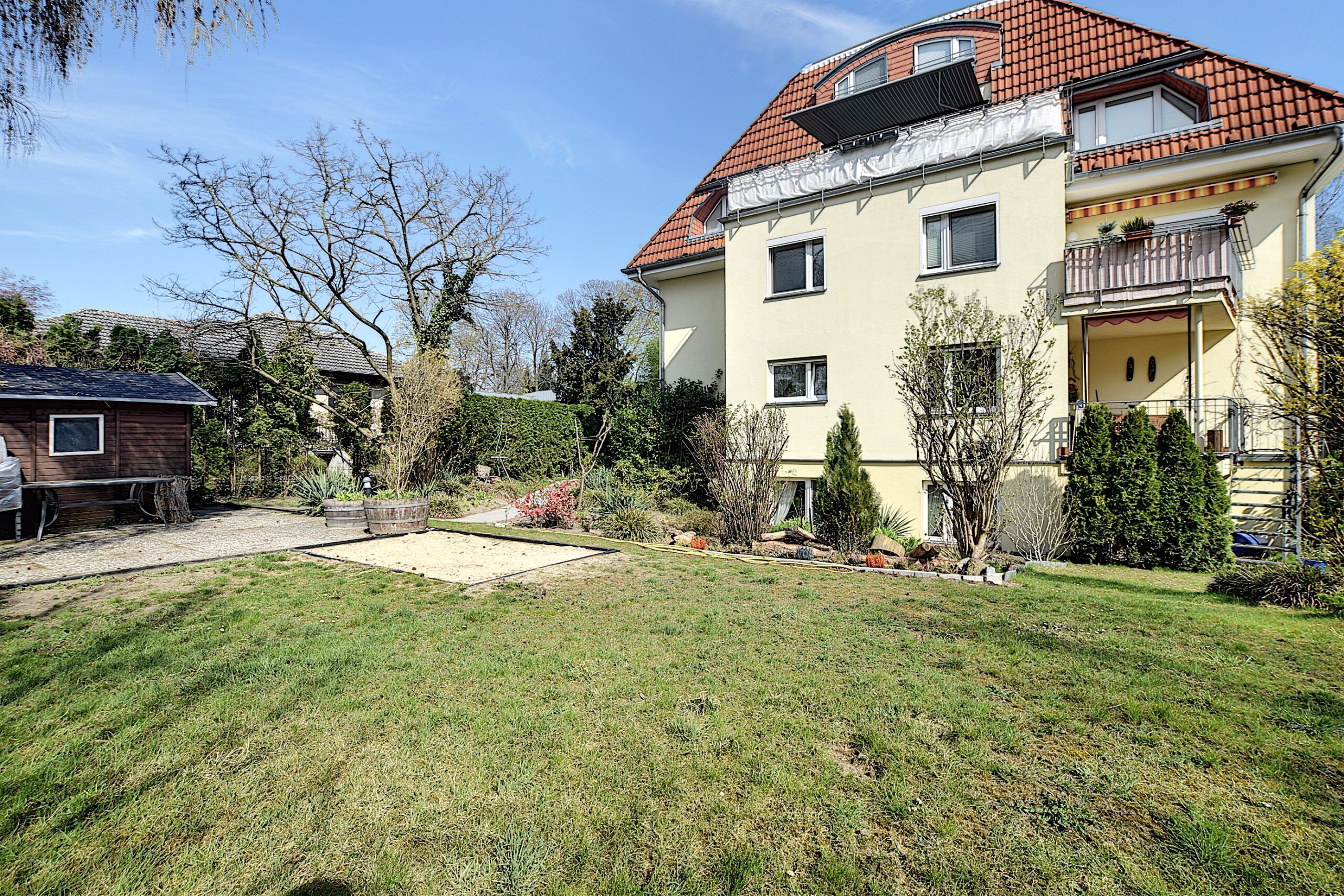 Meergezinswoning met gele pleistergevel, balkon en rode dakpannen, frontaal vanuit tuin met zandbak en houthok