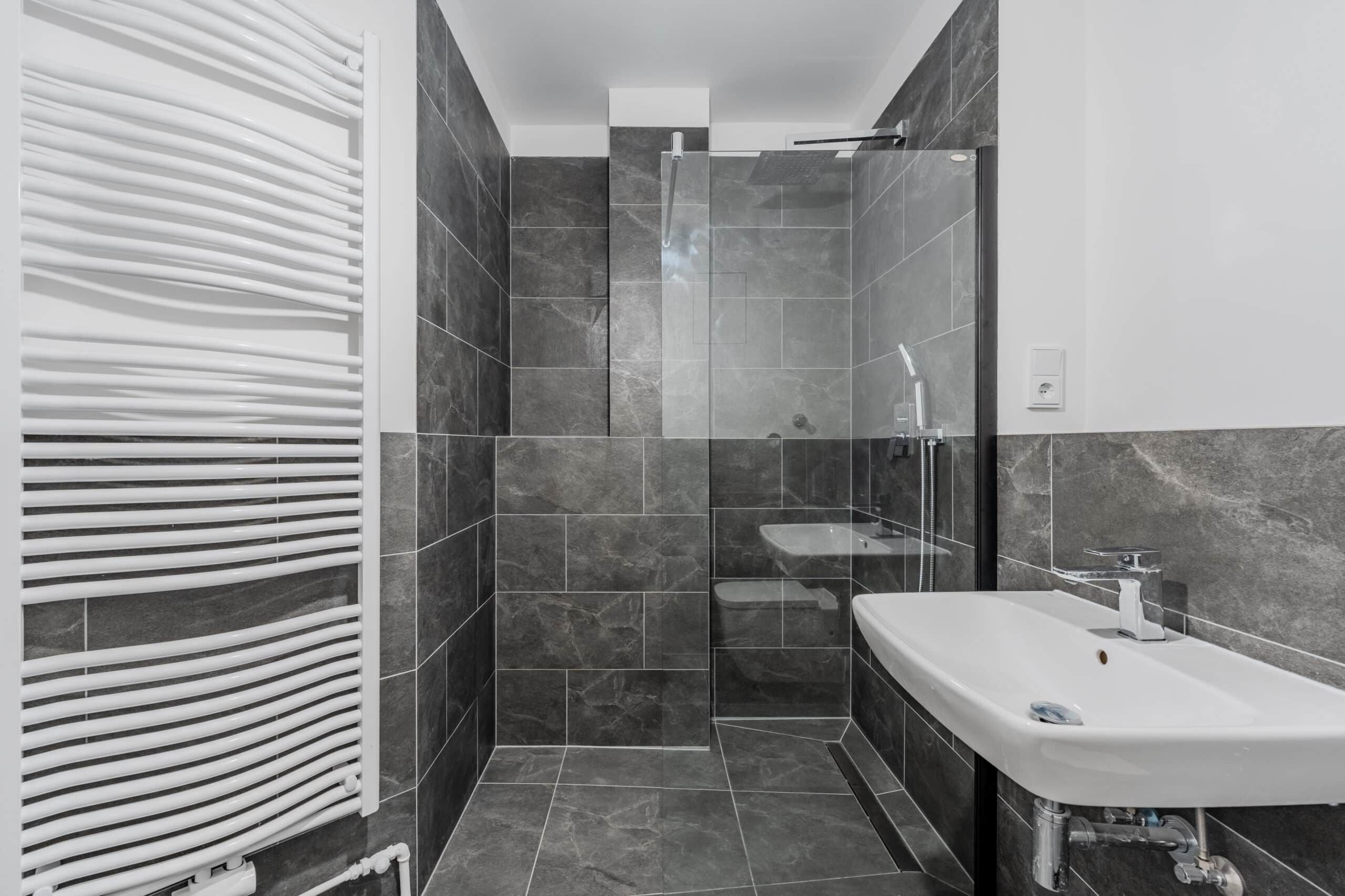Frontansicht modernes Wohnungsbadezimmer mit grauen Fliesen begehbarer Dusche Glaswand Handtuchheizkörper