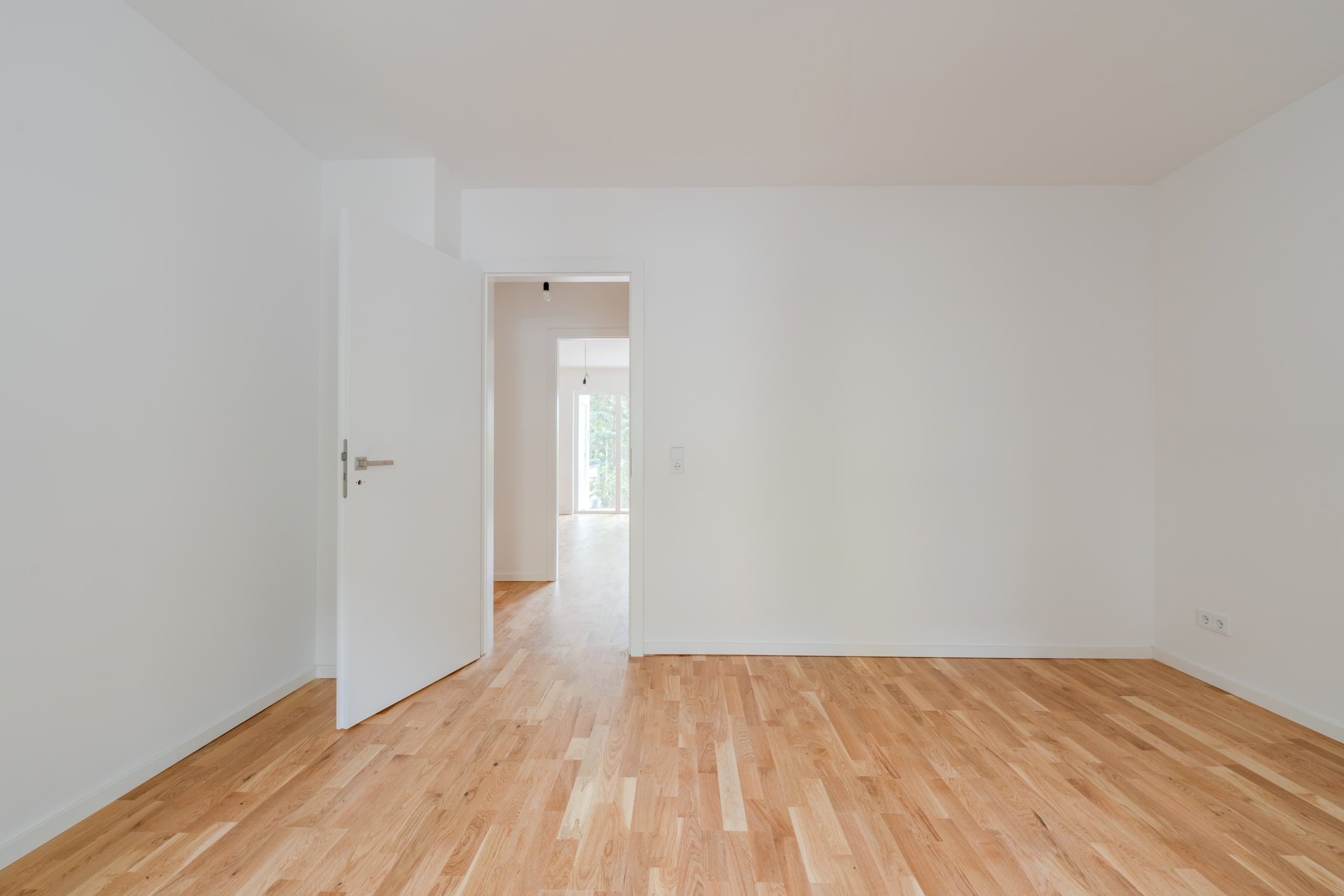 Leeres Zimmer mit hellem Parkettfußboden und offener Tür zur Diele und dahinter sichtbare Glasfront