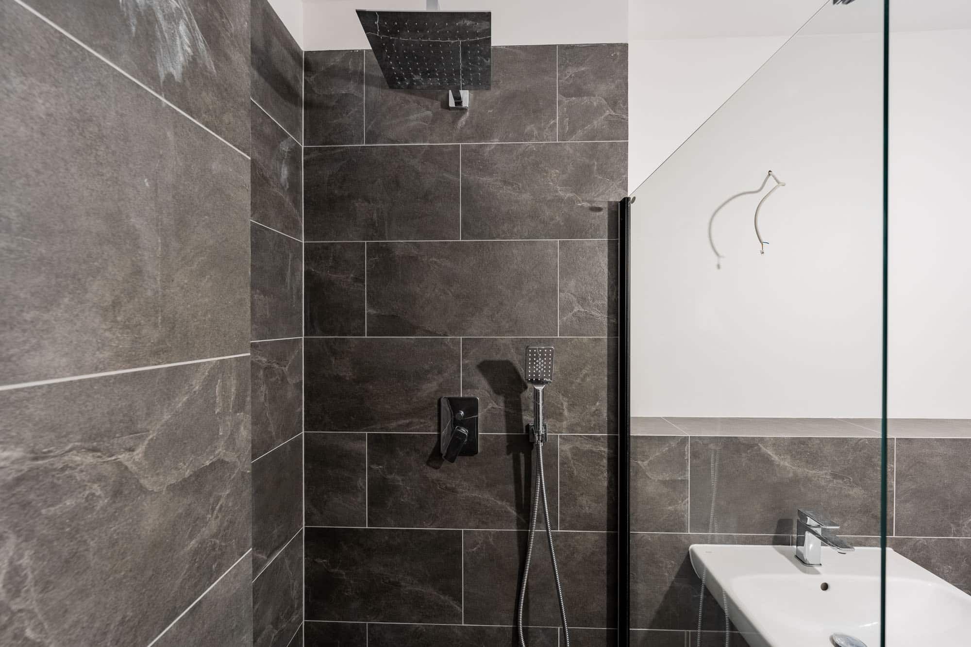 Badezimmer mit moderner Duschkabine in dunkelgrauen Fliesen, Regenduschkopf, Handbrause und weißem Waschbecken