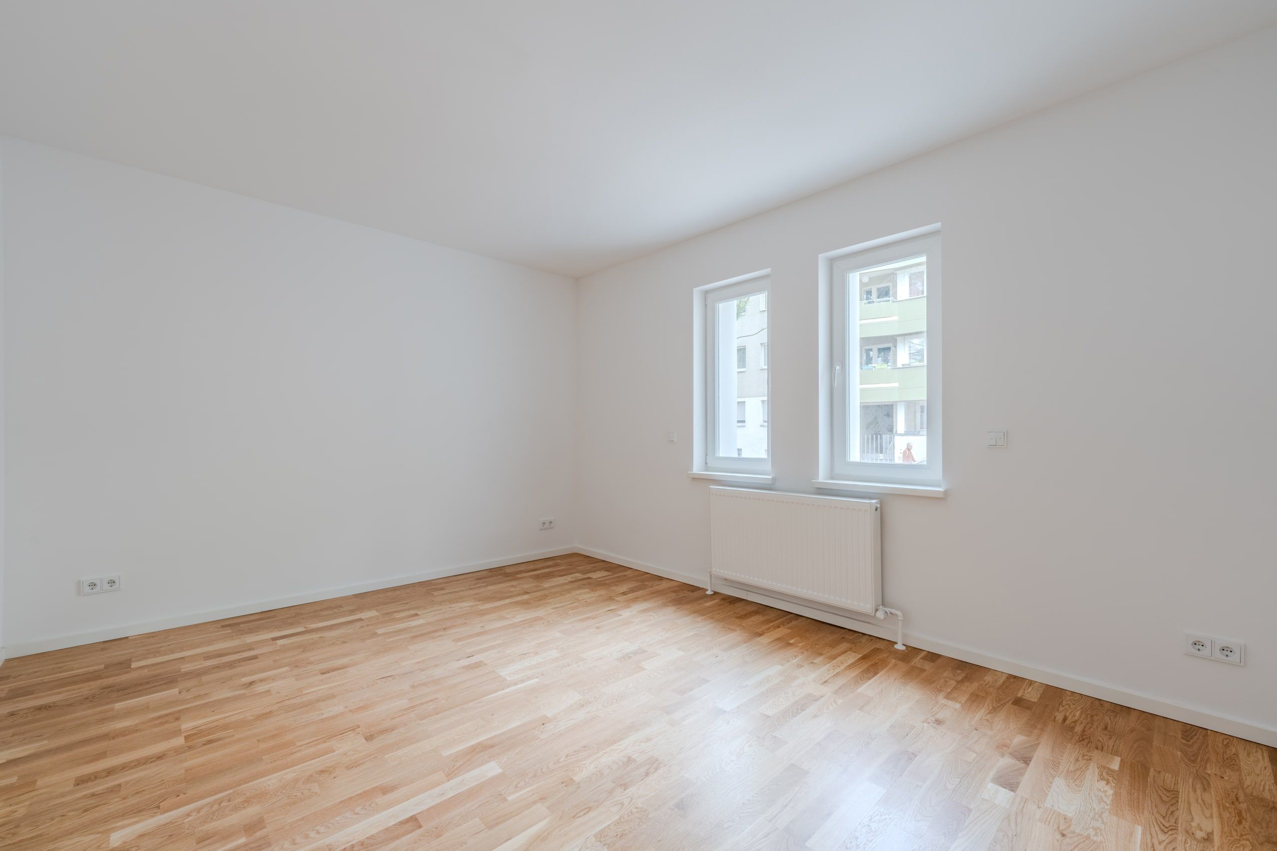 Leerstehendes Wohnungszimmer mit hellem Parkettboden zwei Fenstern und Wandheizung aus Eckperspektive