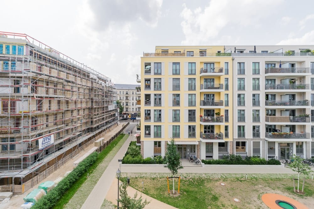 Frontansicht eines modernen Mehrfamilienhauses mit gelb-weißer Fassade, Balkonen und Baugerüst am Nachbargebäude