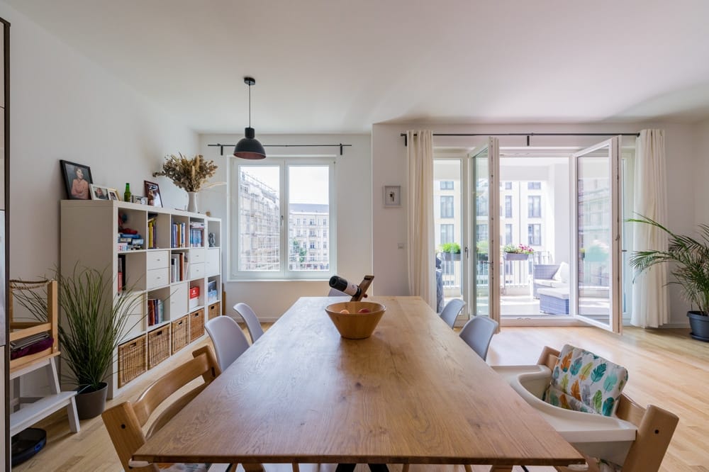 Innenansicht offene Wohnküche moderner Stadtwohnung mit Holztisch, Regalwand und bodentiefen Fenstertüren zum Balkon