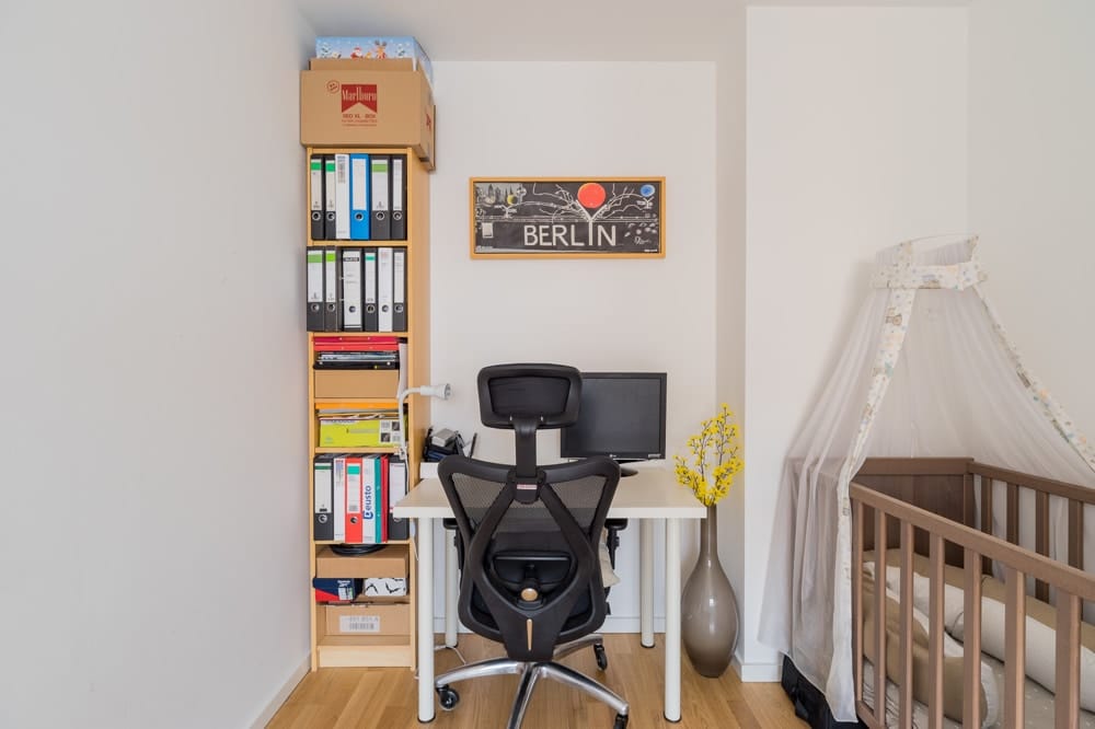 Wohnungsnische als Homeoffice mit weißem Schreibtisch, schwarzem Bürostuhl, Regalwand mit Ordnern und Babybett