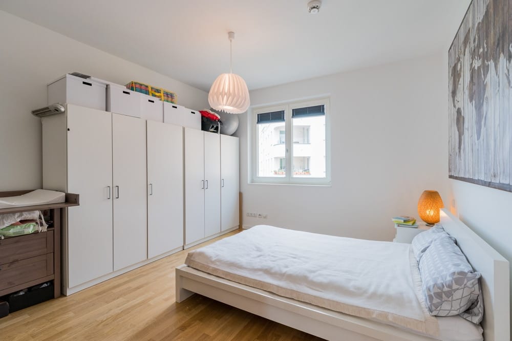 Innenansicht eines Schlafzimmers mit Doppelbett, großem weißen Kleiderschrank, Parkettboden und Fenster mit Balkonblick