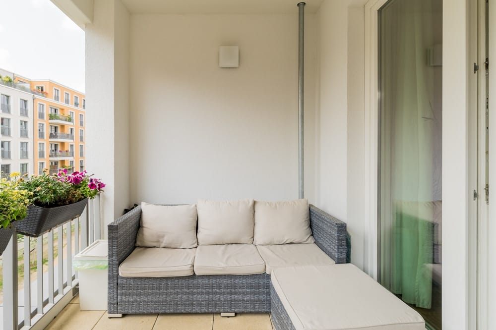 Moderner Balkon einer Wohnung mit grauem Rattansofa, Polsterauflagen, Blumenkästen am Geländer, hell verputzte Fassade