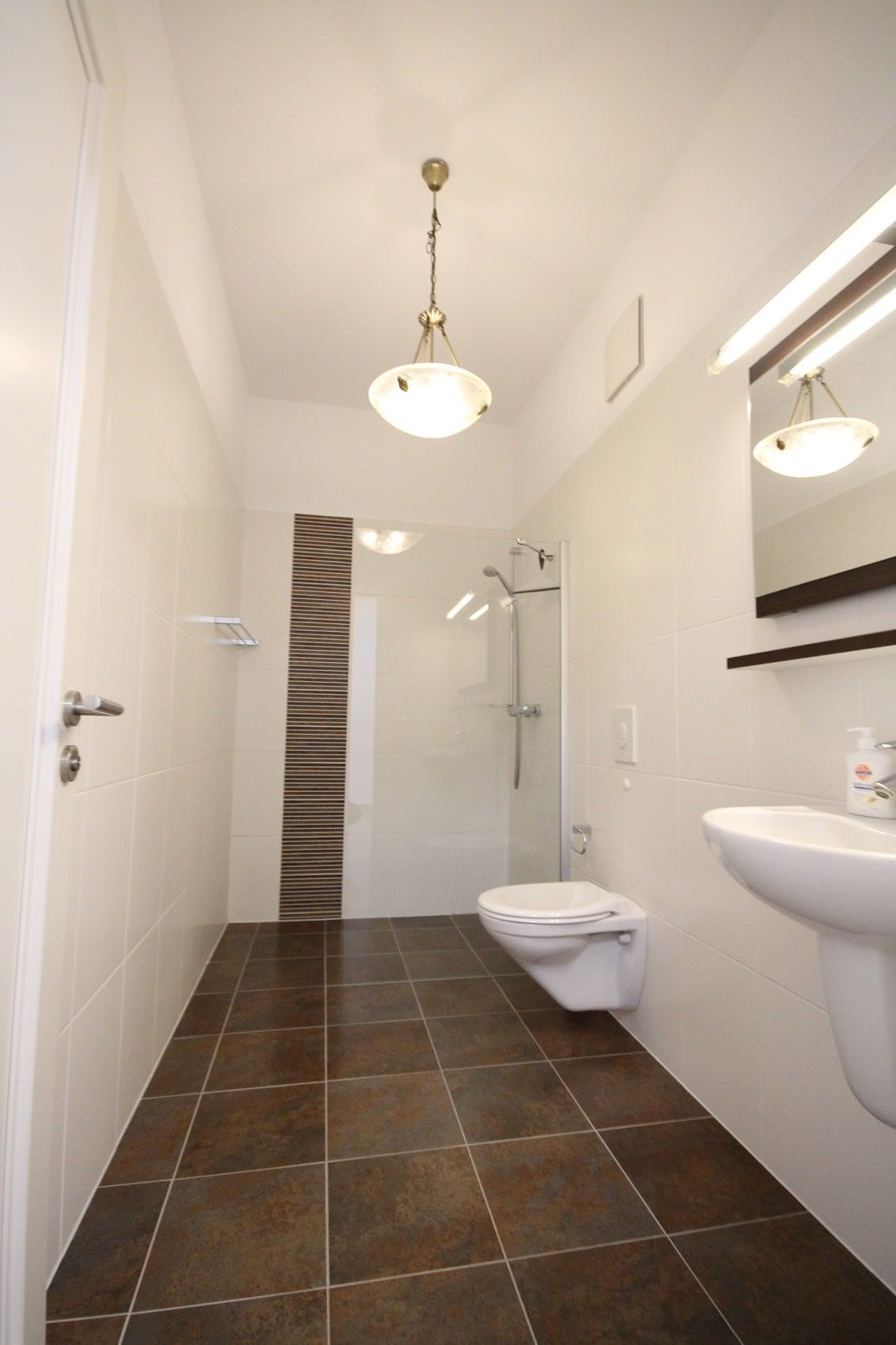 Interno di un bagno con cabina doccia, parete divisoria in vetro, WC sospeso, lavabo e piastrelle marroni a pavimento