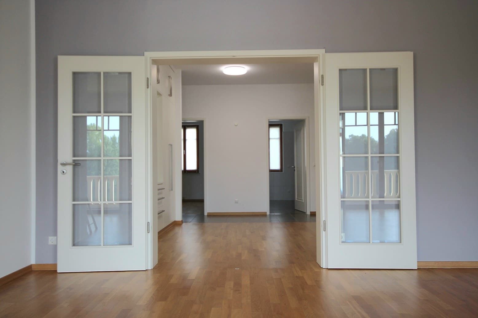 Interno di un soggiorno vuoto con pavimento in parquet, porte in vetro bianco alla francese e vista su un luminoso corridoio con finestre