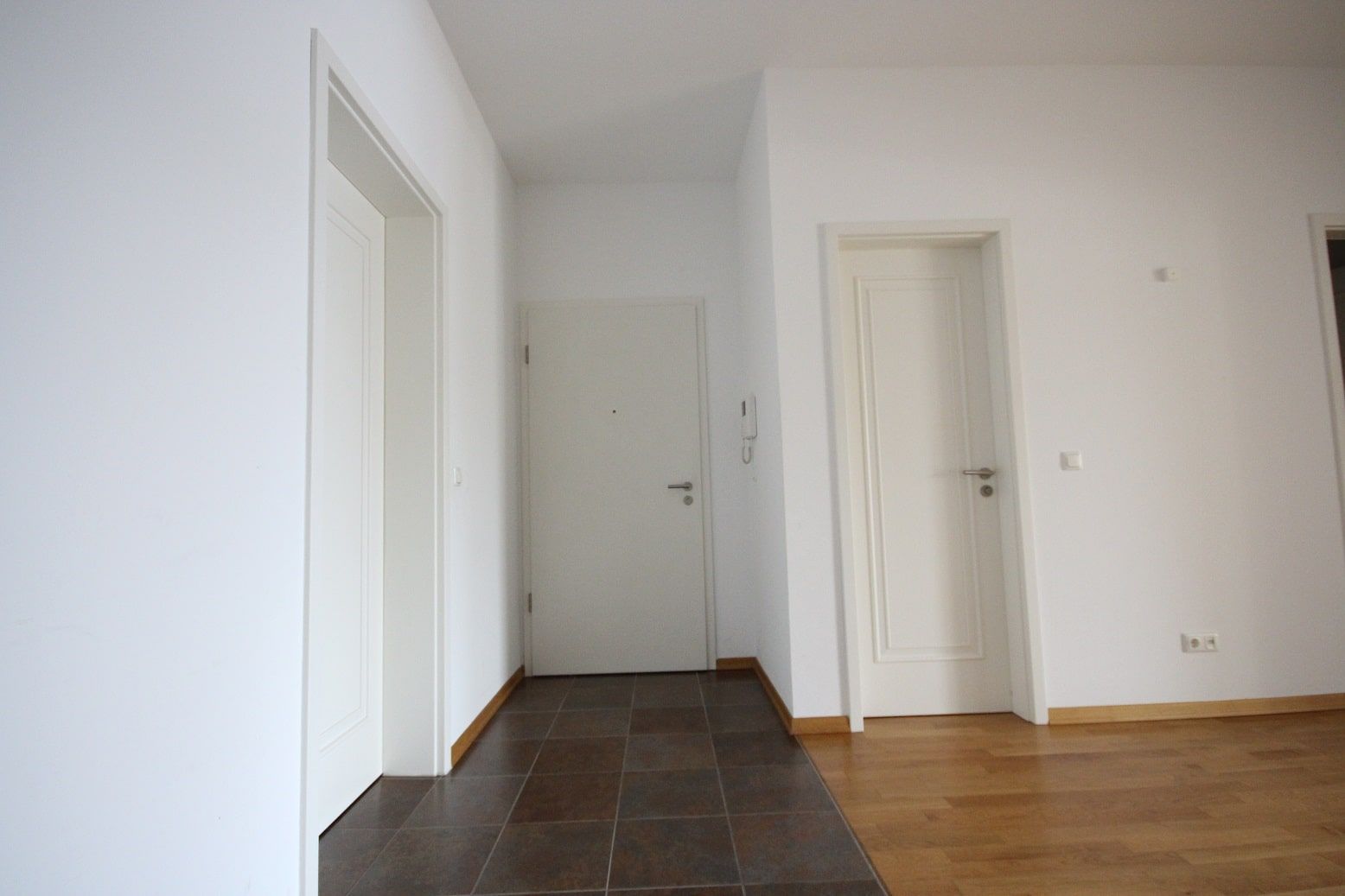 Interno di un appartamento di nuova costruzione, visto di fronte, con tre porte bianche, pavimento in piastrelle marroni e parquet