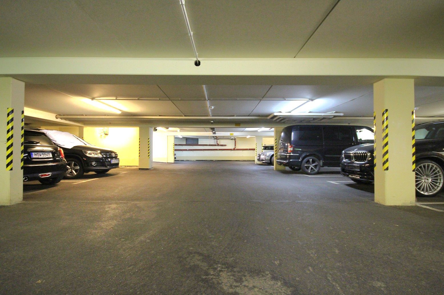 Garage sotterraneo ben illuminato, con pilastri in cemento, posti auto contrassegnati e diverse auto parcheggiate, visto da una prospettiva intermedia
