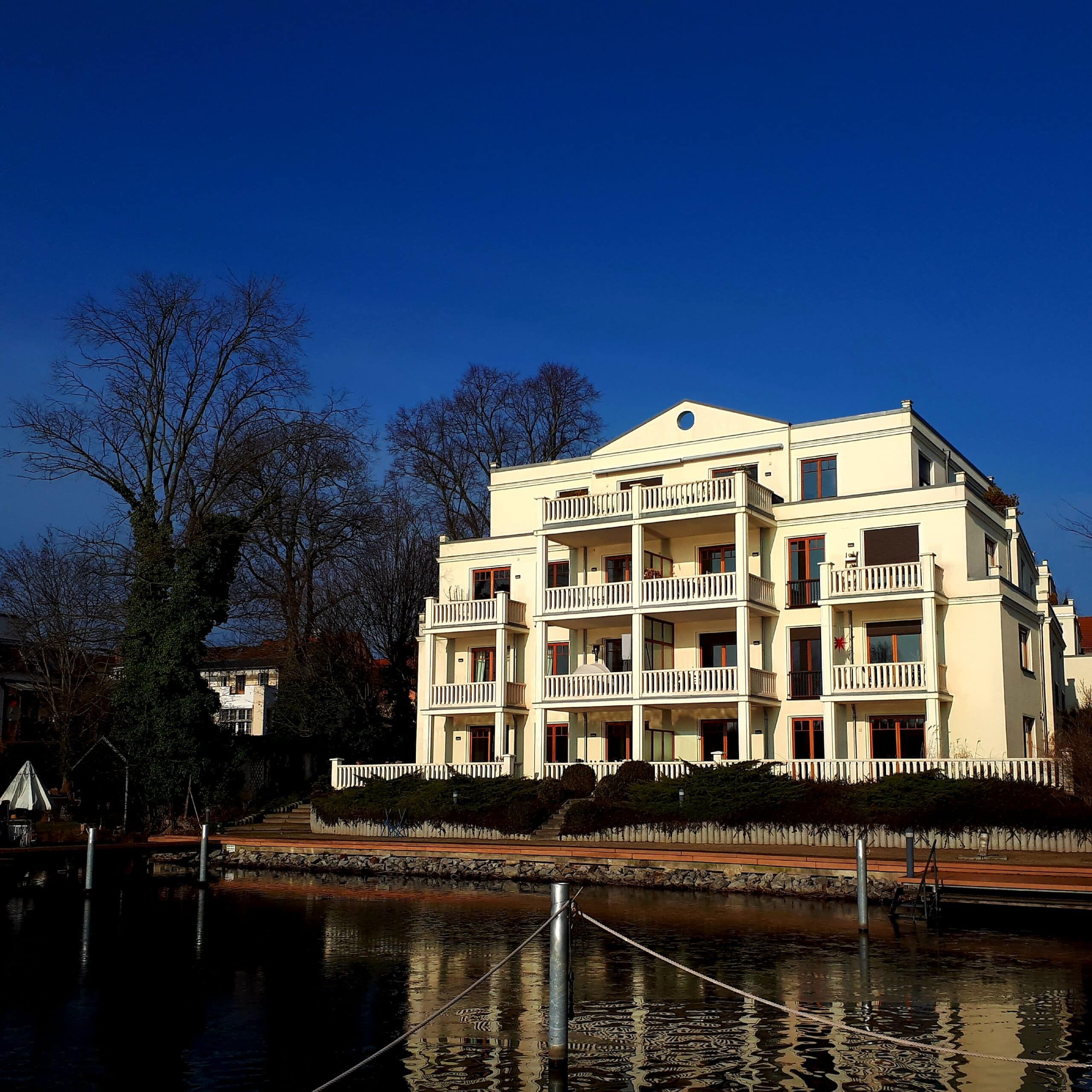 Vista frontale di un classico condominio con facciata intonacata di bianco, tre balconi sovrapposti e la riva del fiume