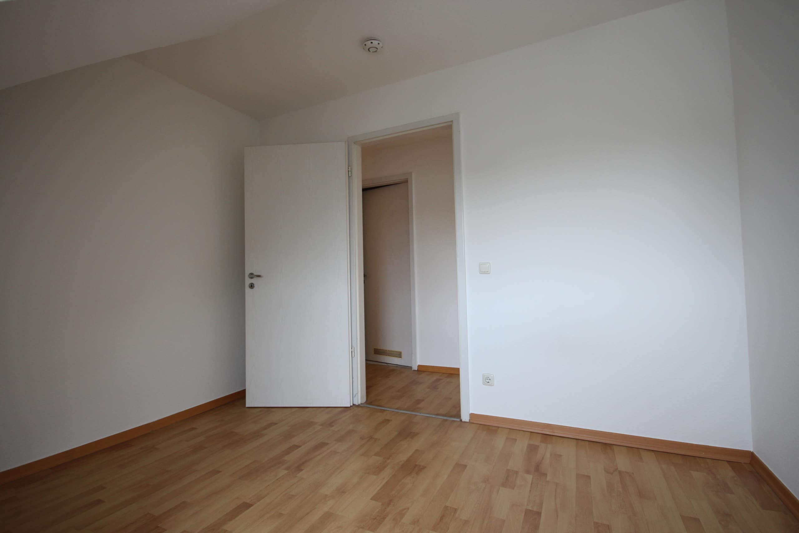 Leer stehender Wohnraum mit Laminatboden und weißen Wänden, Blick auf offene Zimmertür zum Flur