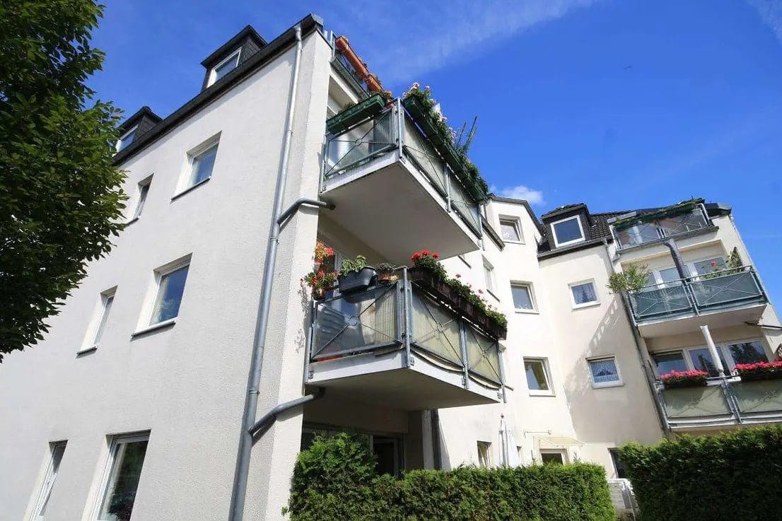 Blick von unten auf mehrgeschossiges Wohnhaus mit weißen Putzfassaden und Balkonen mit Glasgeländer und Pflanzen