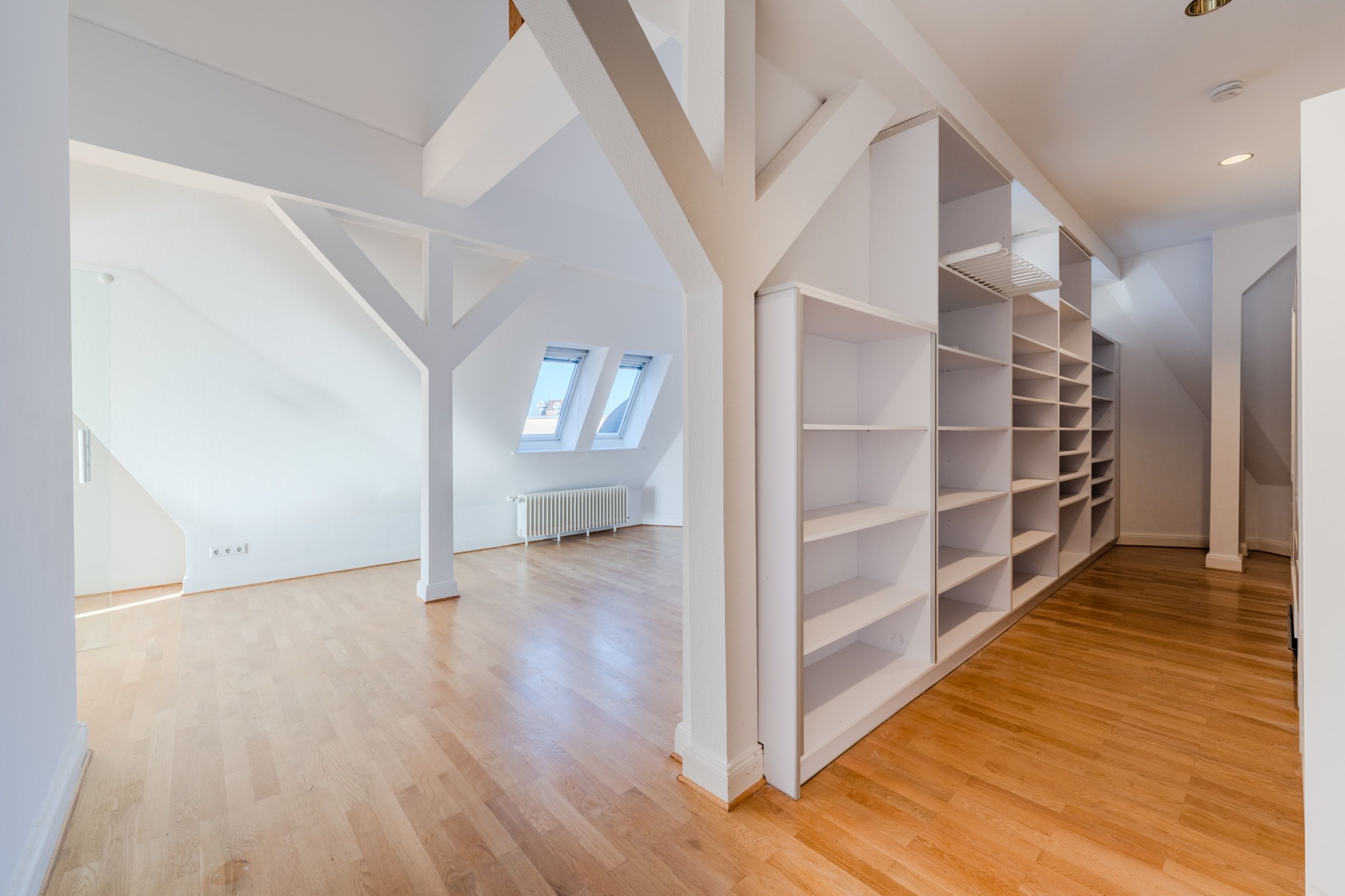 Vista interna di una mansarda ristrutturata con travi a vista bianche, pavimento in parquet, finestre Velux e scaffali a incasso