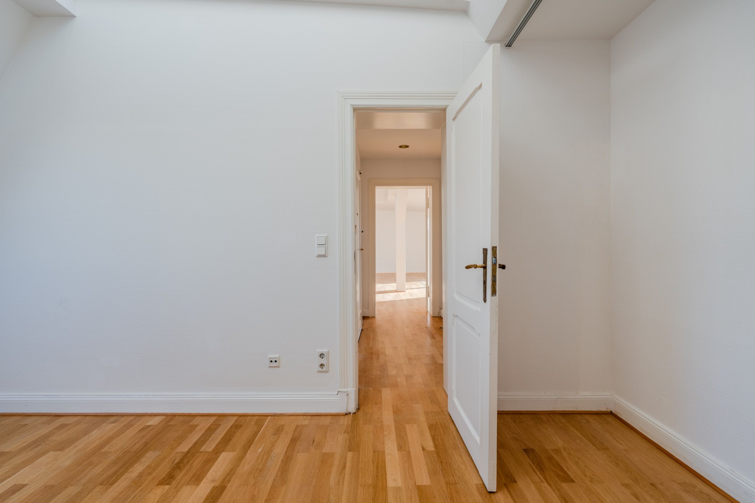 Interno di un appartamento in un edificio storico con pavimento in parquet oliato, pareti bianche e porta aperta che dà sul luminoso corridoio