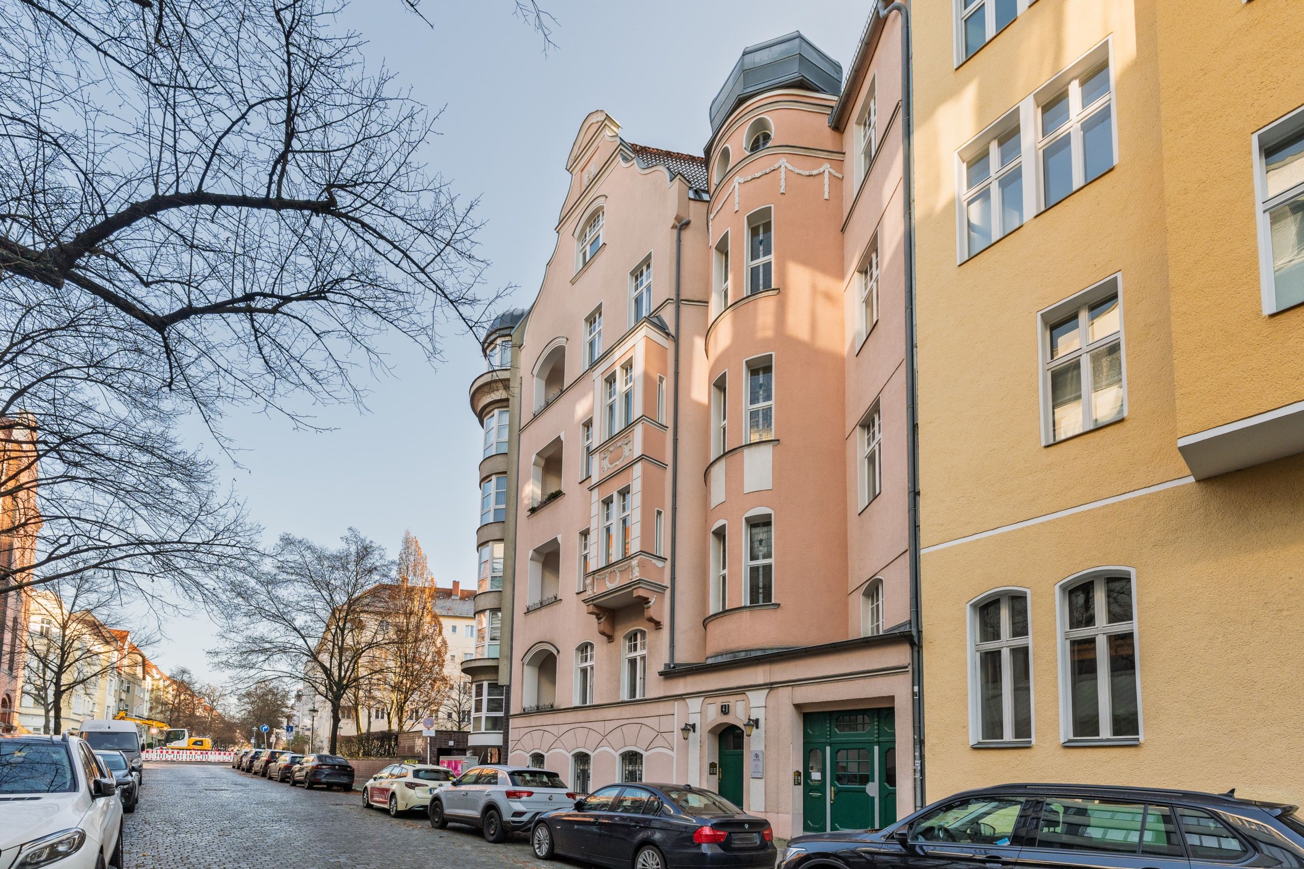 Prospettiva dalla strada di una facciata di un edificio residenziale in stile storicista, di colore rosa pastello, con bovindi, balconi e porta d’ingresso verde