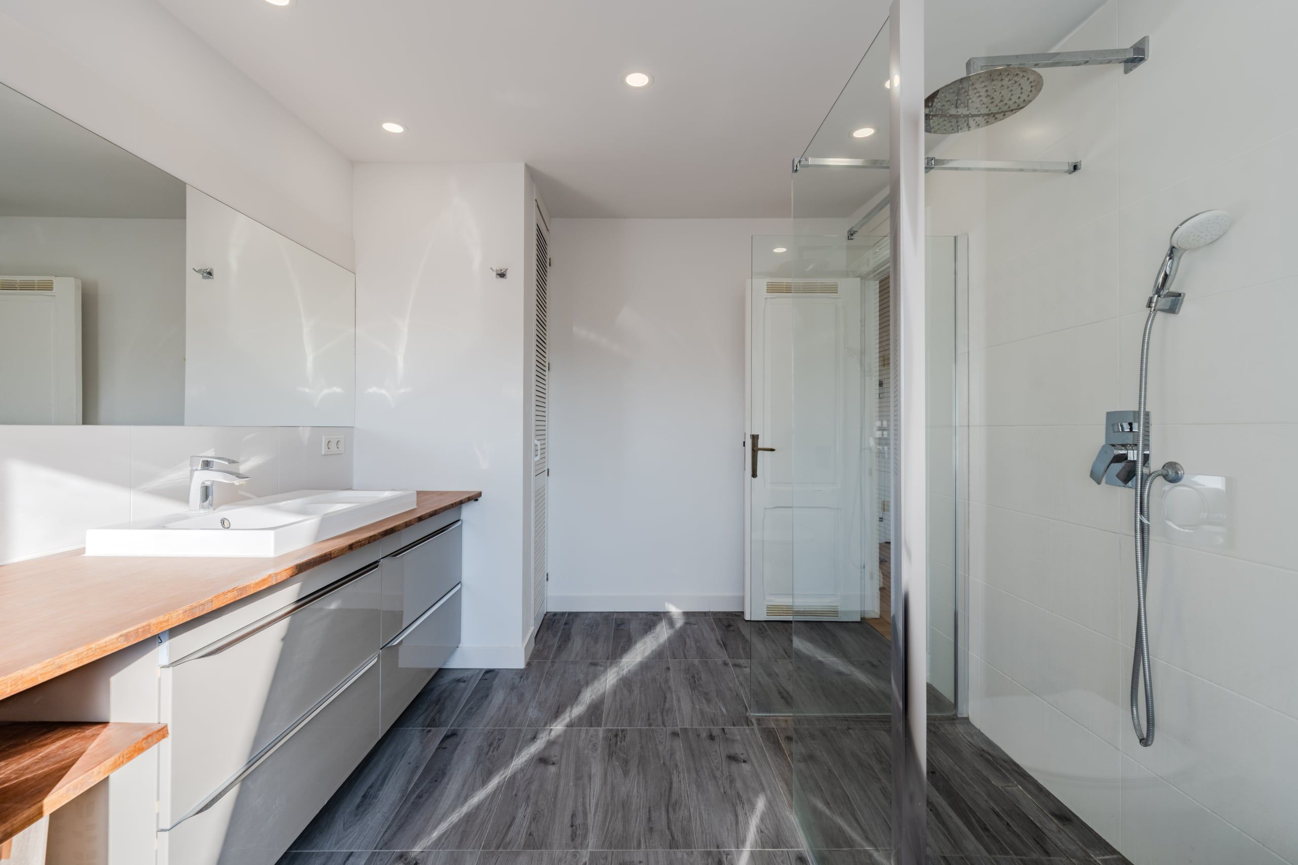 Vista interna di un bagno moderno con cabina doccia in vetro, doppio lavabo e piano di lavoro in legno, con la porta in primo piano