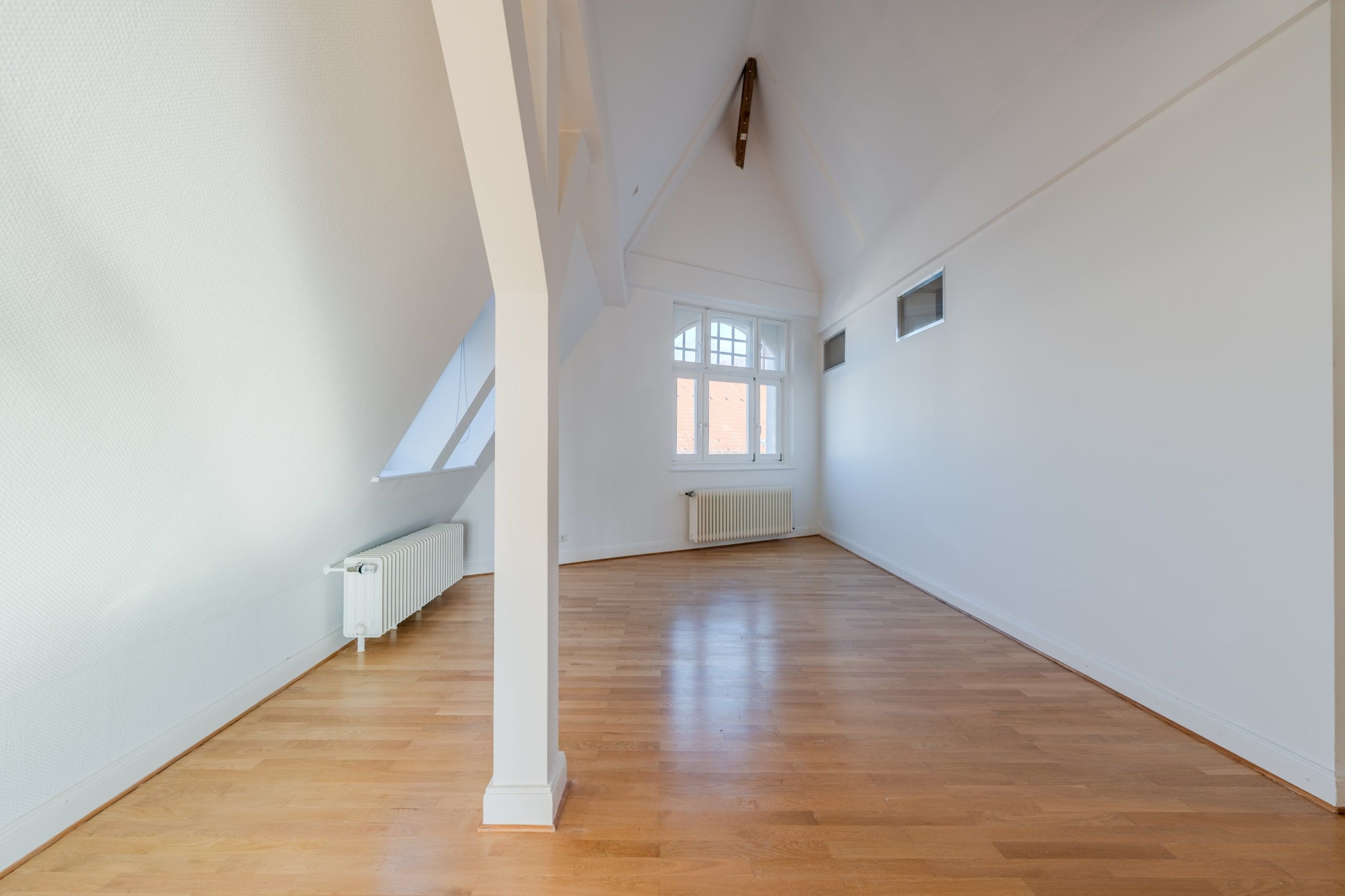 sala vuota nell'attico con pareti bianche e pavimento in legno, ripresa con grandangolo; si vedono la pendenza del tetto e i termosifoni