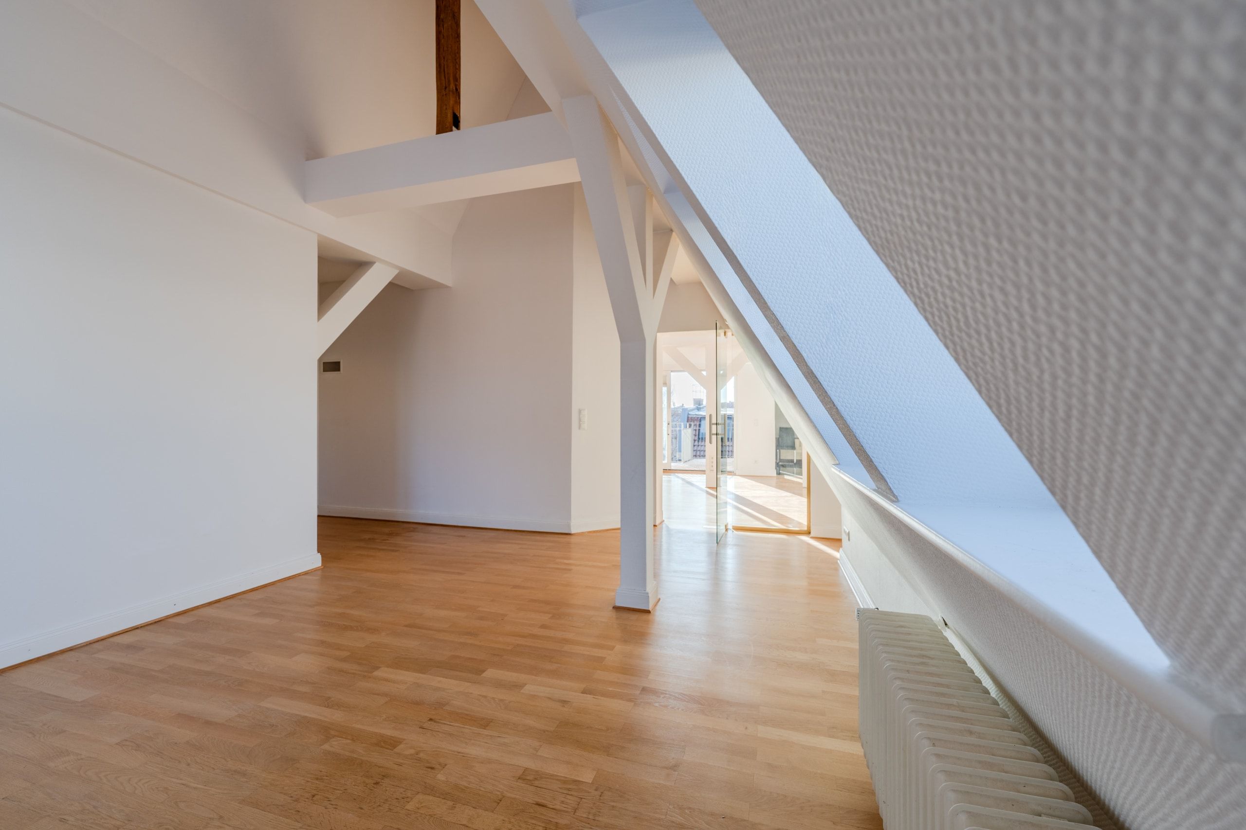 Sala aperta nell'attico con travi a vista, pavimento in parquet, finestra da tetto inclinata, radiatore e porta a vetri a tutta altezza che dà sul balcone