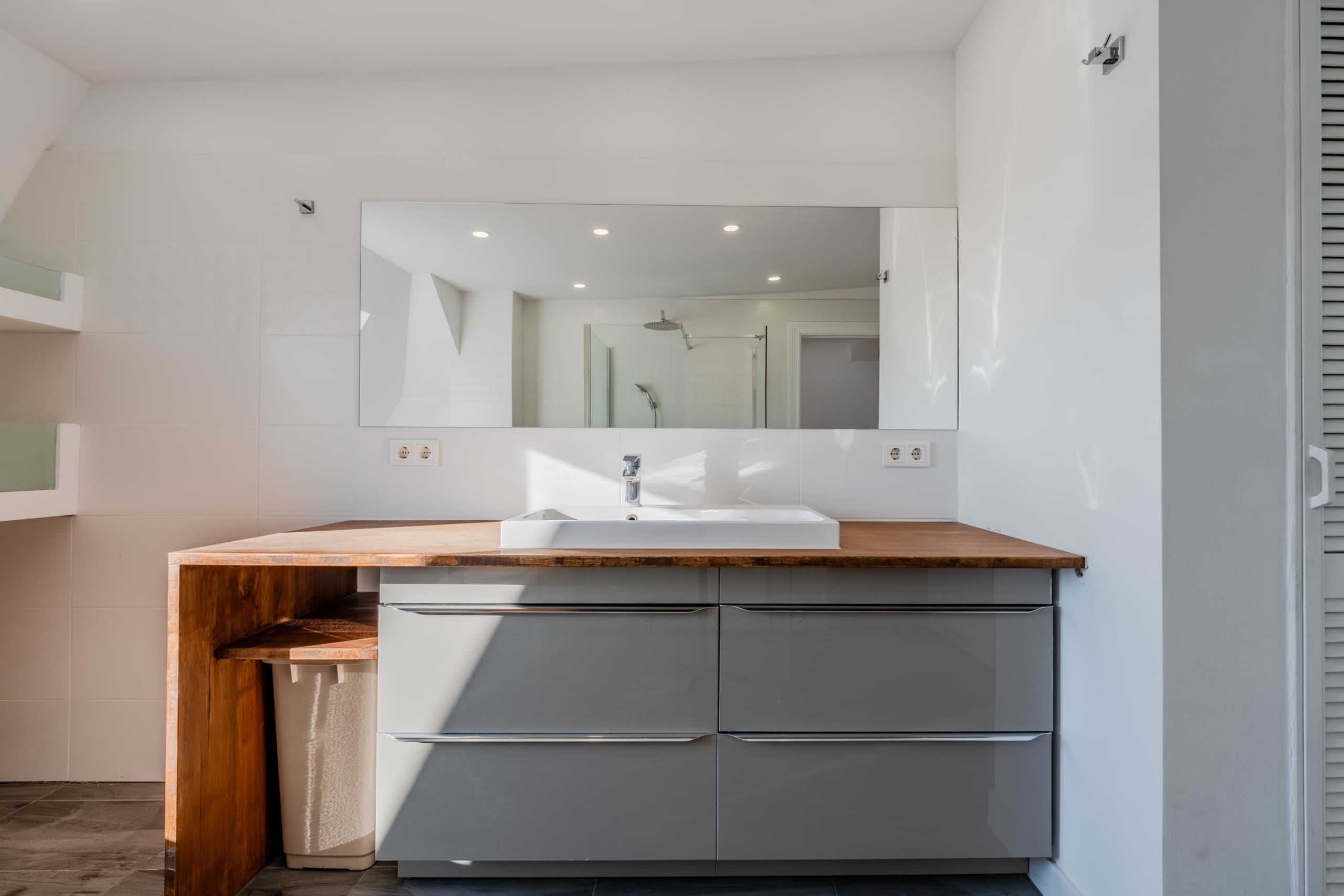 Foto degli interni di un bagno moderno con mobile lavabo grigio e piano di lavoro in legno; specchio e doccia sullo sfondo