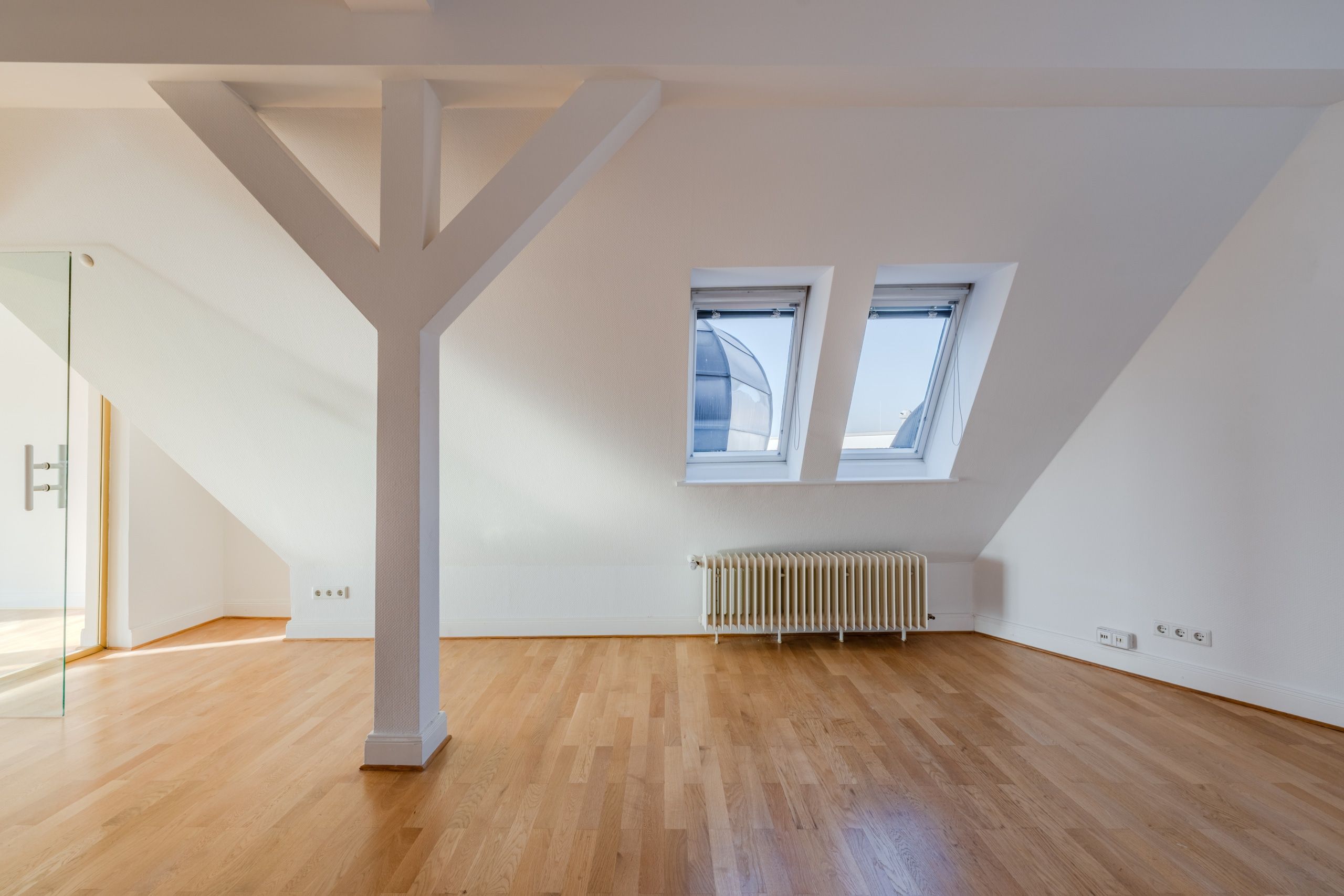 Vista interna di una mansarda vuota con pavimento in parquet, soffitto spiovente, due lucernari, un pilastro centrale e un termosifone