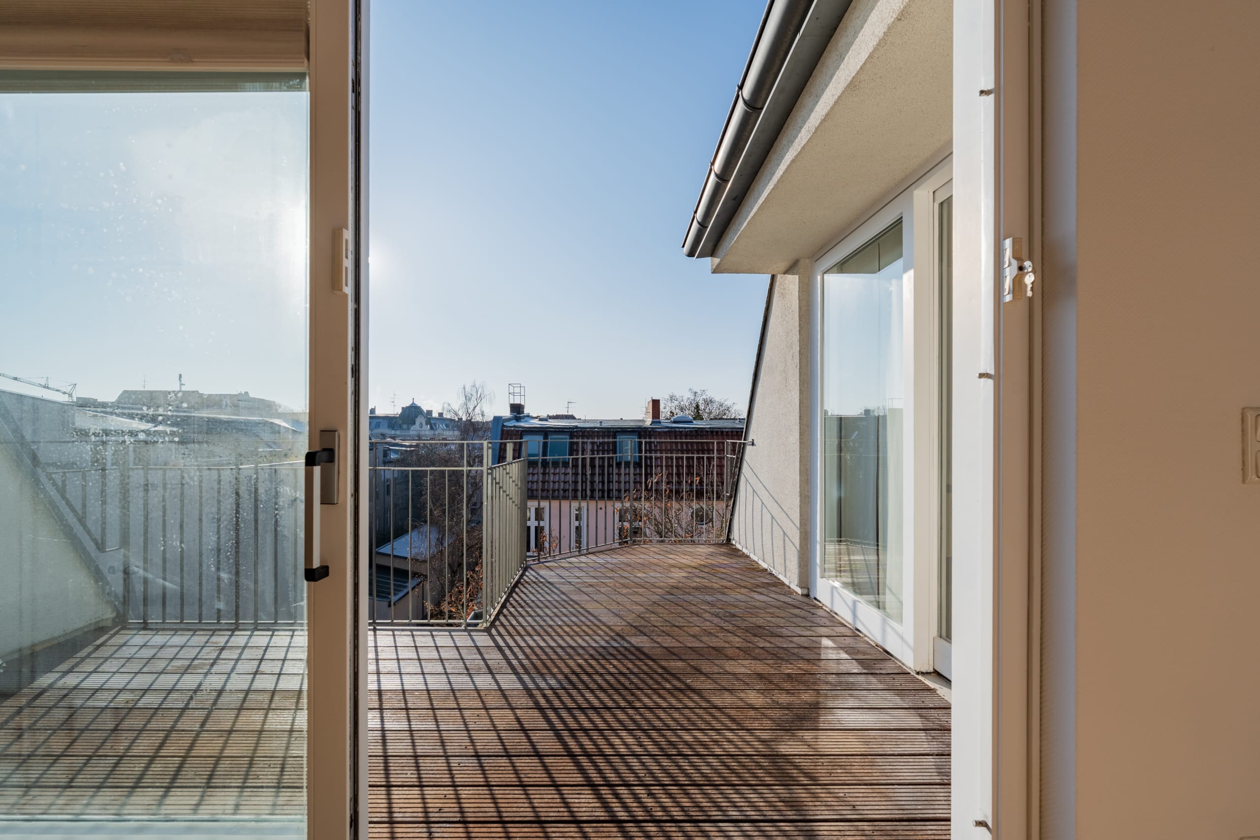 Balcone di un appartamento mansardato, accessibile dall'interno tramite una porta scorrevole aperta, con terrazza in legno e ringhiera in metallo, con vista sui tetti della città