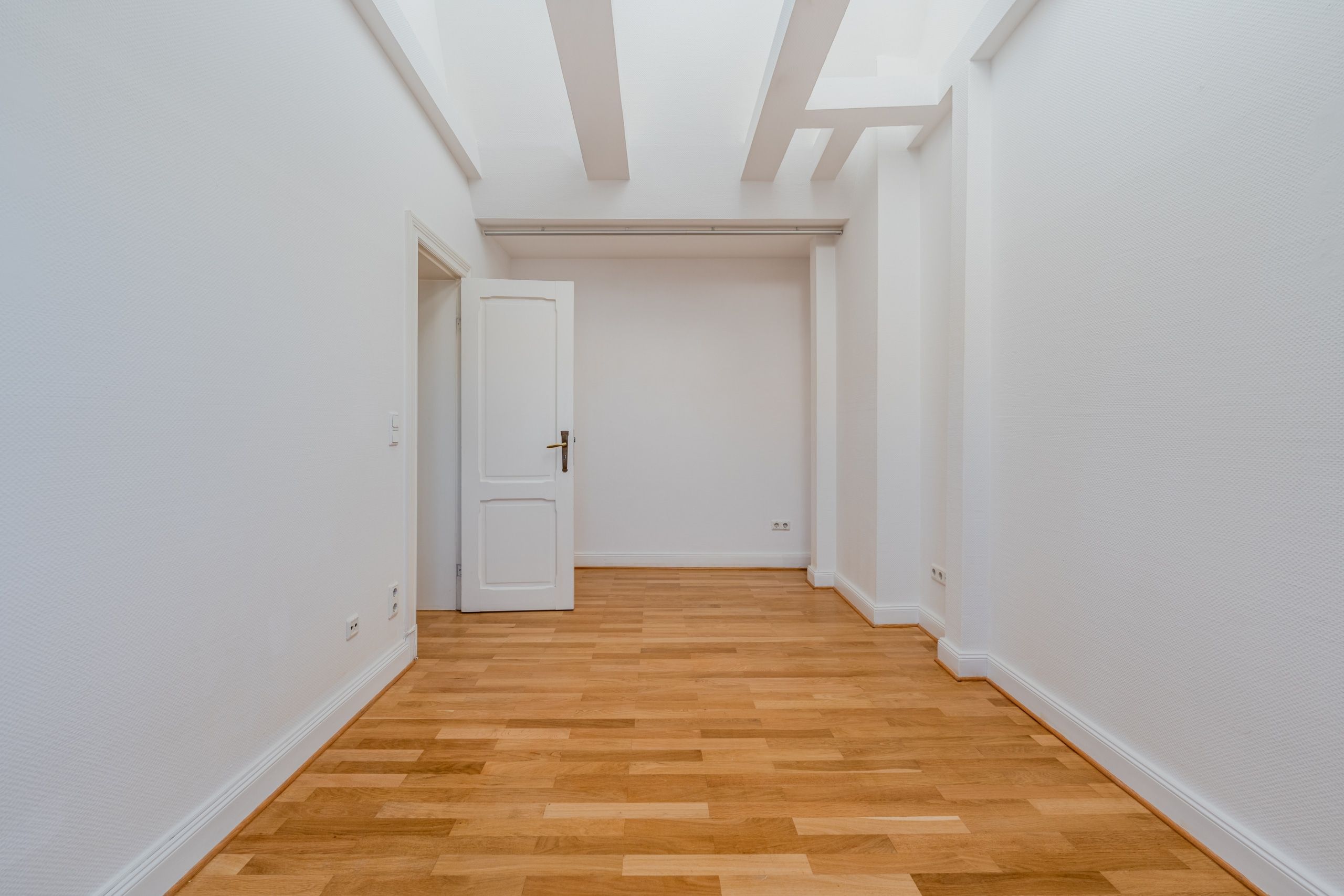 Vista interna del corridoio di un appartamento vuoto con la porta aperta, pavimento in parquet di legno e travi a vista bianche