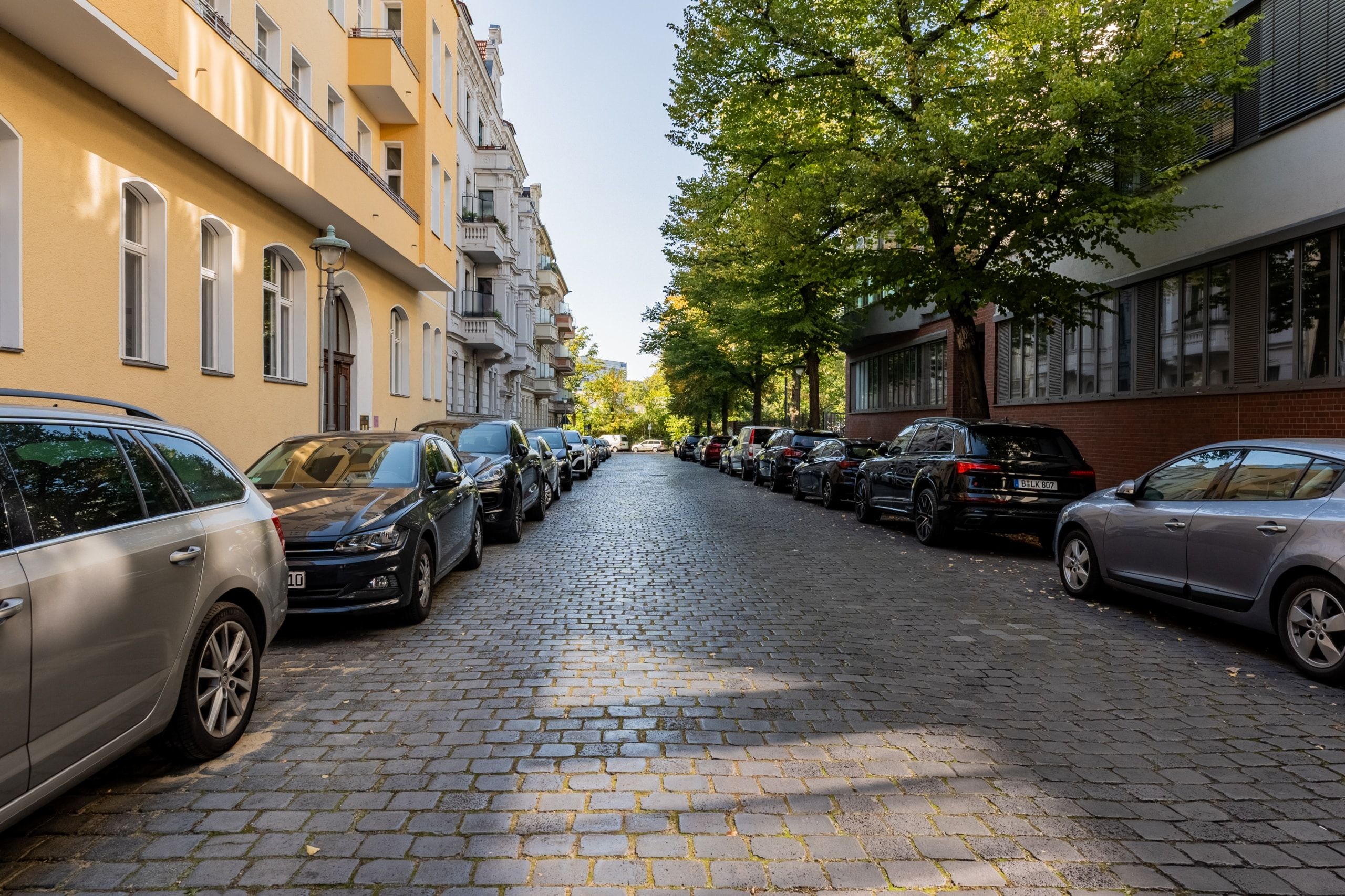 Strada residenziale lastricata con file di auto parcheggiate tra un vecchio edificio giallo e un edificio moderno, vista dalla strada
