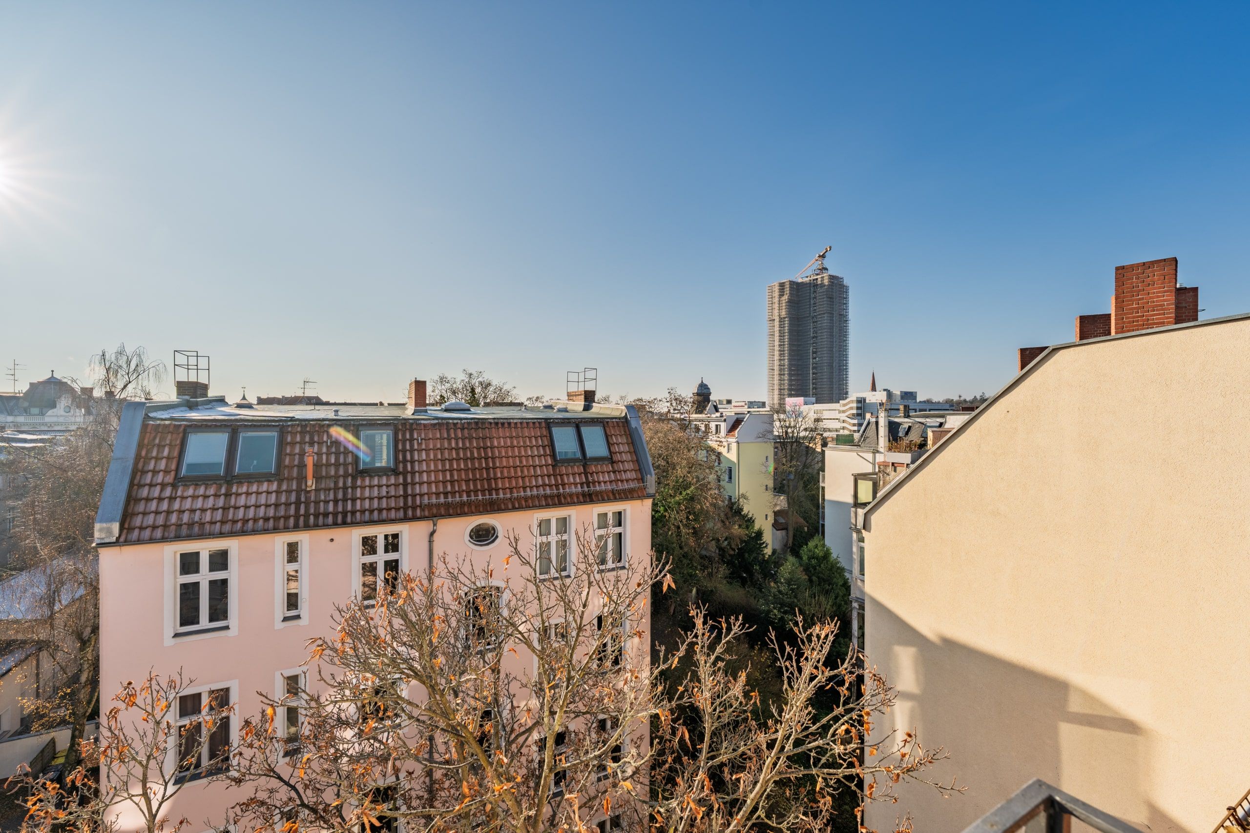Vista dall’alto di un vecchio edificio residenziale a tre piani con tetto ad abbaino e un albero spoglio in autunno, davanti al cantiere di un grattacielo