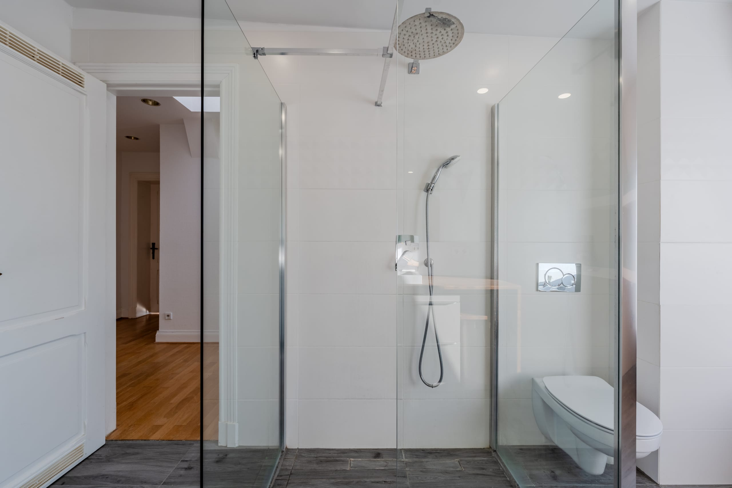 Vista interna di un bagno moderno con cabina doccia in vetro a filo pavimento, soffione a pioggia e doccetta, WC sospeso