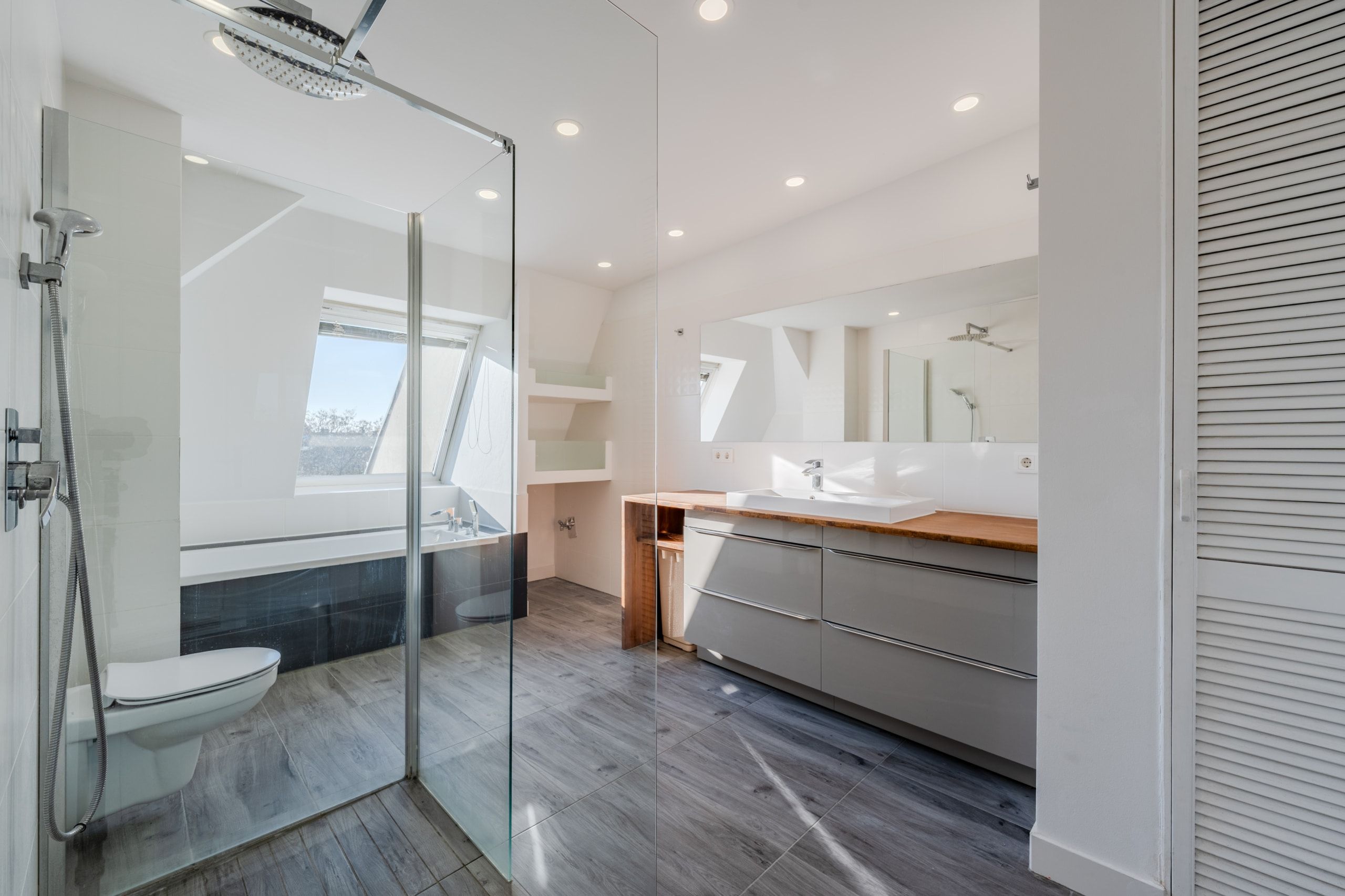 Bagno mansardato con cabina doccia in vetro, vasca da bagno sotto la finestra mansardata e doppio lavabo con ripiano in legno