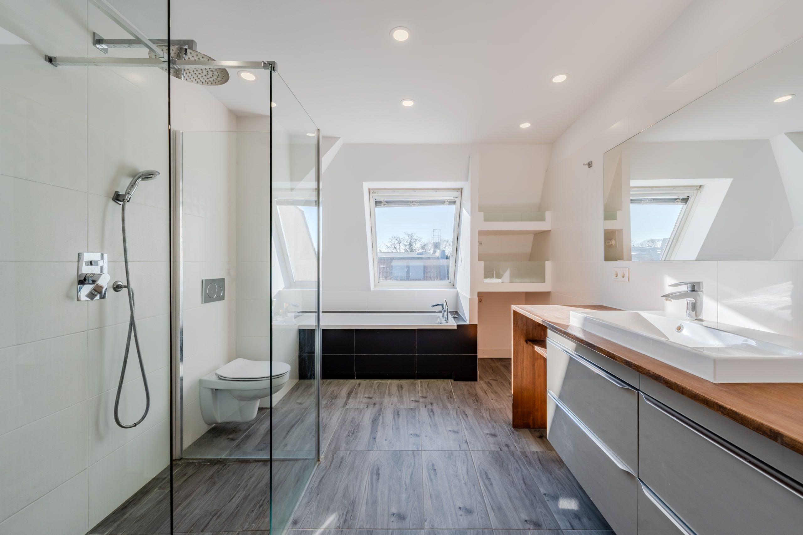 Bagno mansardato in un edificio di nuova costruzione con doccia walk-in, vasca da bagno indipendente sotto la finestra mansardata e lavabo in legno con specchio