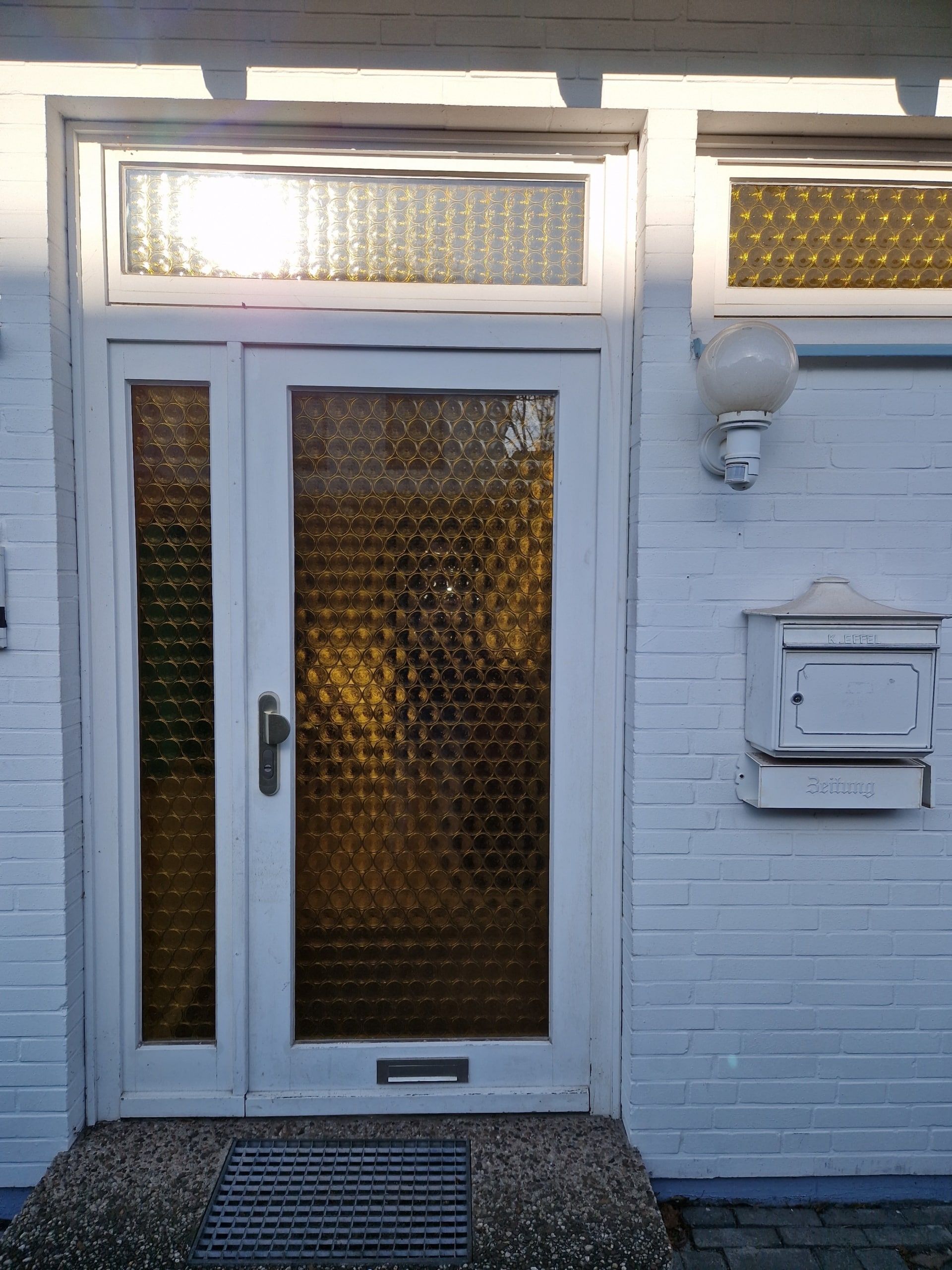 Frontansicht des Eingangsbereichs eines weißen Klinkerhauses mit weißer Tür und gelb getöntem Glaseinsatz neben Briefkasten
