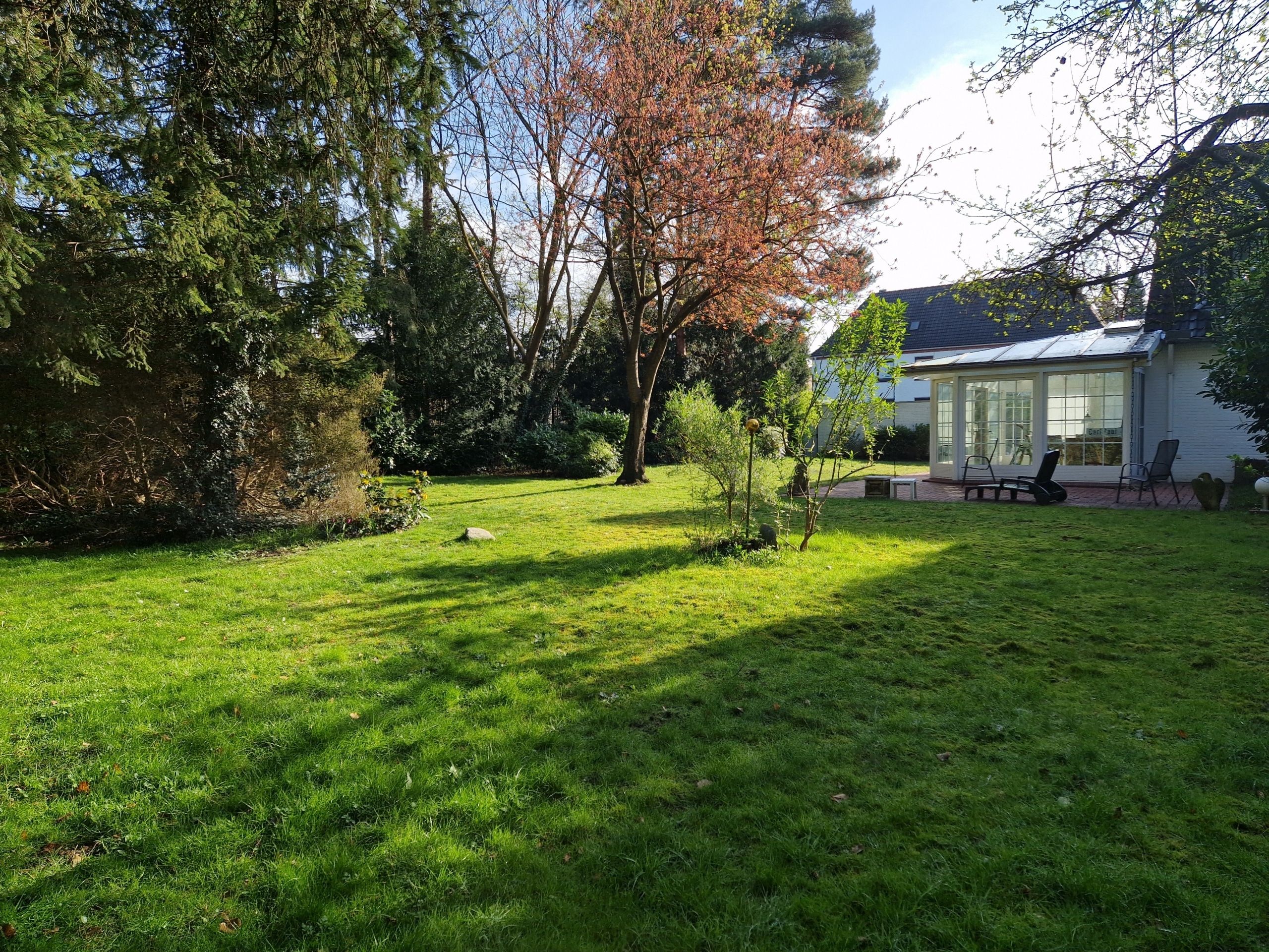 Blick auf Einfamilienhaus mit Wintergarten und Terrassenbereich, davor weitläufiger Rasen und umgebende Bäume