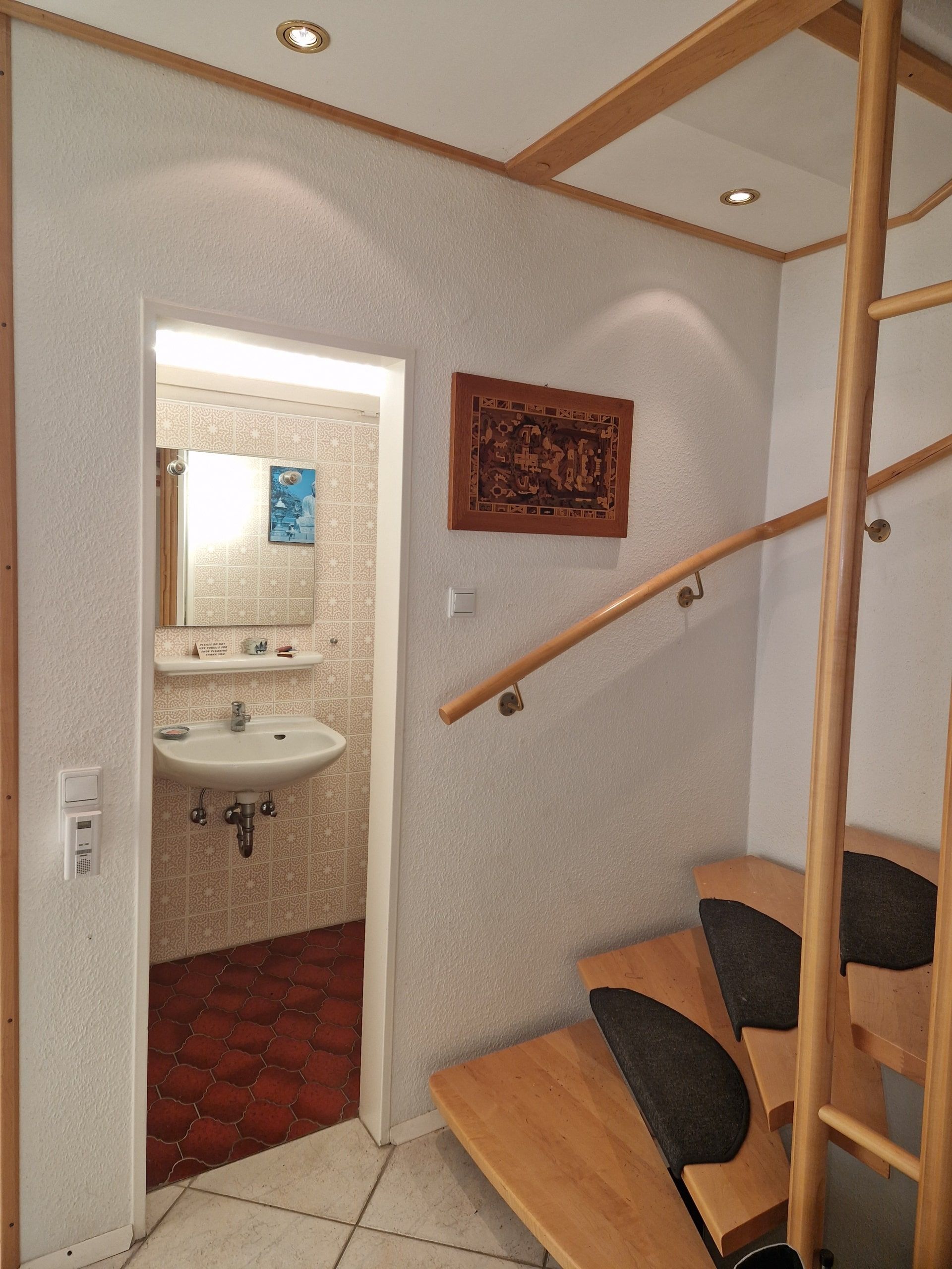 Innenansicht eines Flurs mit Holz-Spindeltreppe und Zugang zu kleinem Badezimmer mit Wandfliesen und Waschbecken