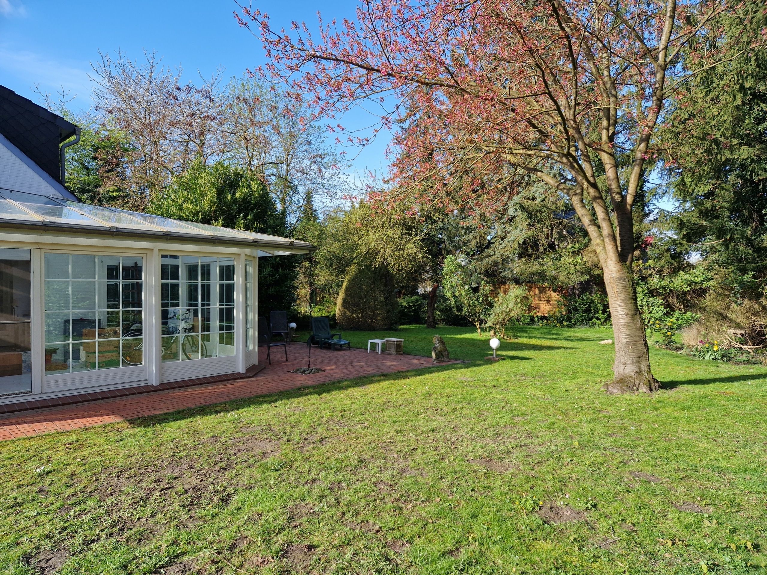 Gartenansicht Einfamilienhaus mit verglastem Wintergarten, Klinkerterrasse, Rasen und blühendem Baum