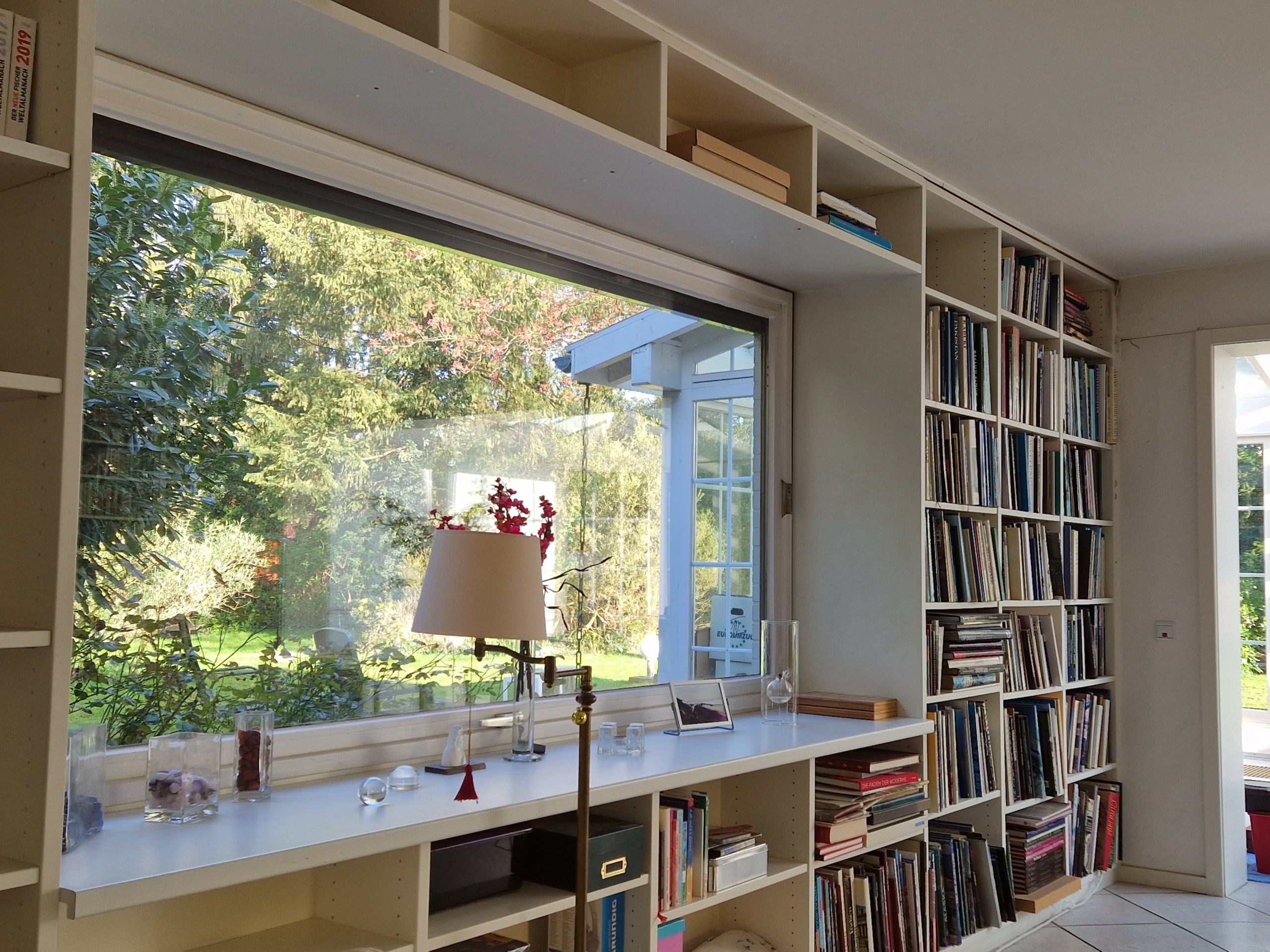 Innenraum eines Wohnhauses mit bodentiefer Fensterfront Blick in Garten raumhohe Bücherregale und Fensterbank mit Lampe