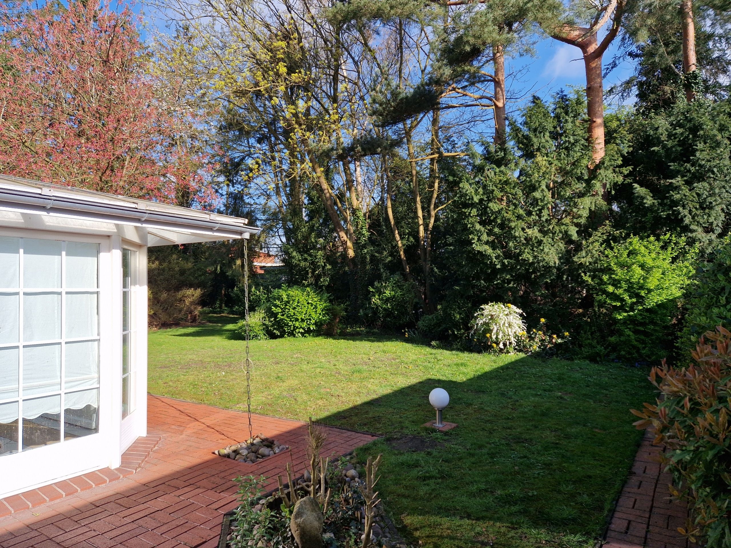 Blick von roter Klinker-Terrasse auf weißen Wintergarten und grünen Garten mit Rasen und Bäumen