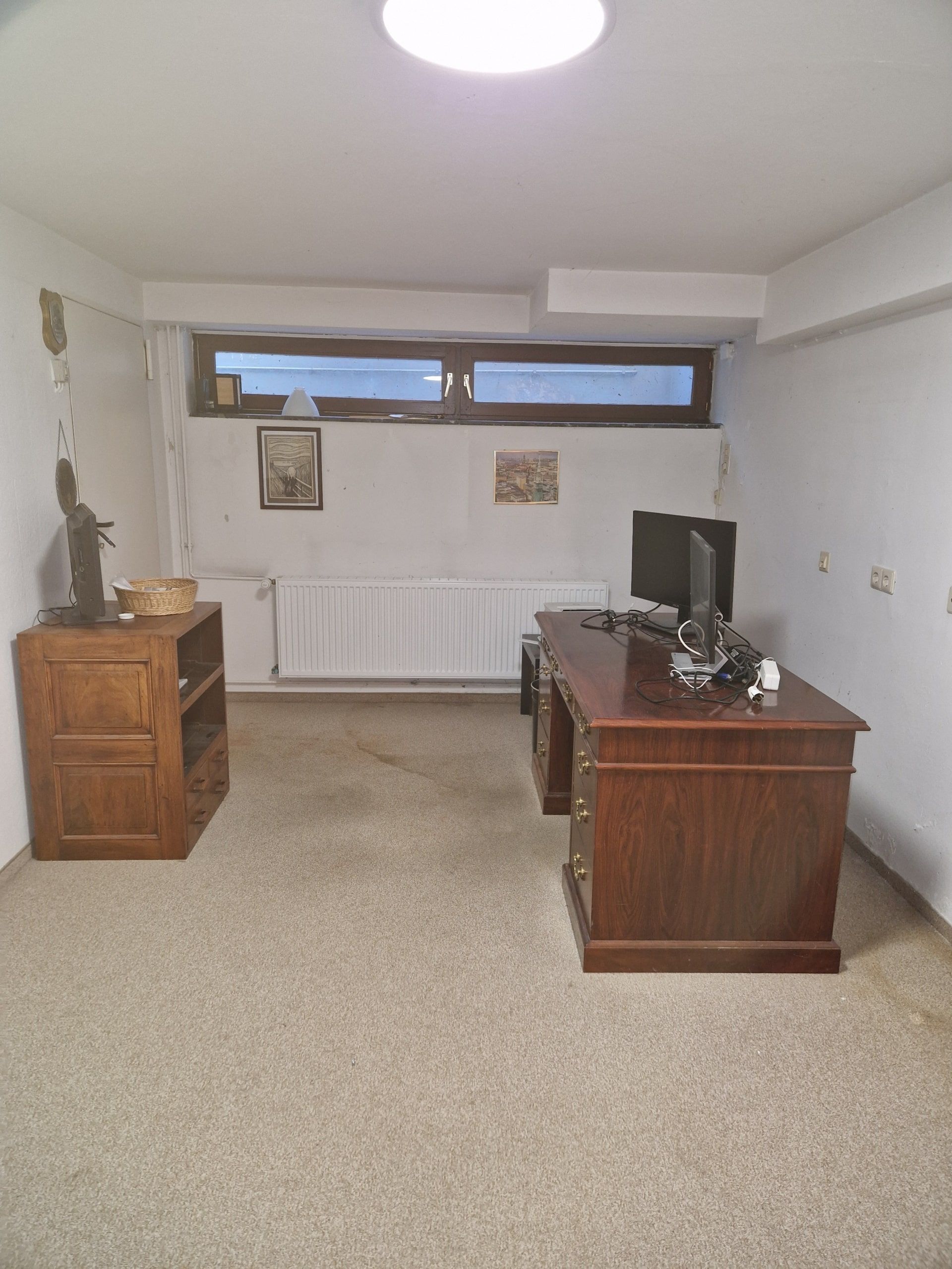 heller kellerraum mit niedrigem hochkantfenster von seitlicher perspektive antiker dunkler holzschreibtisch monitor und regal