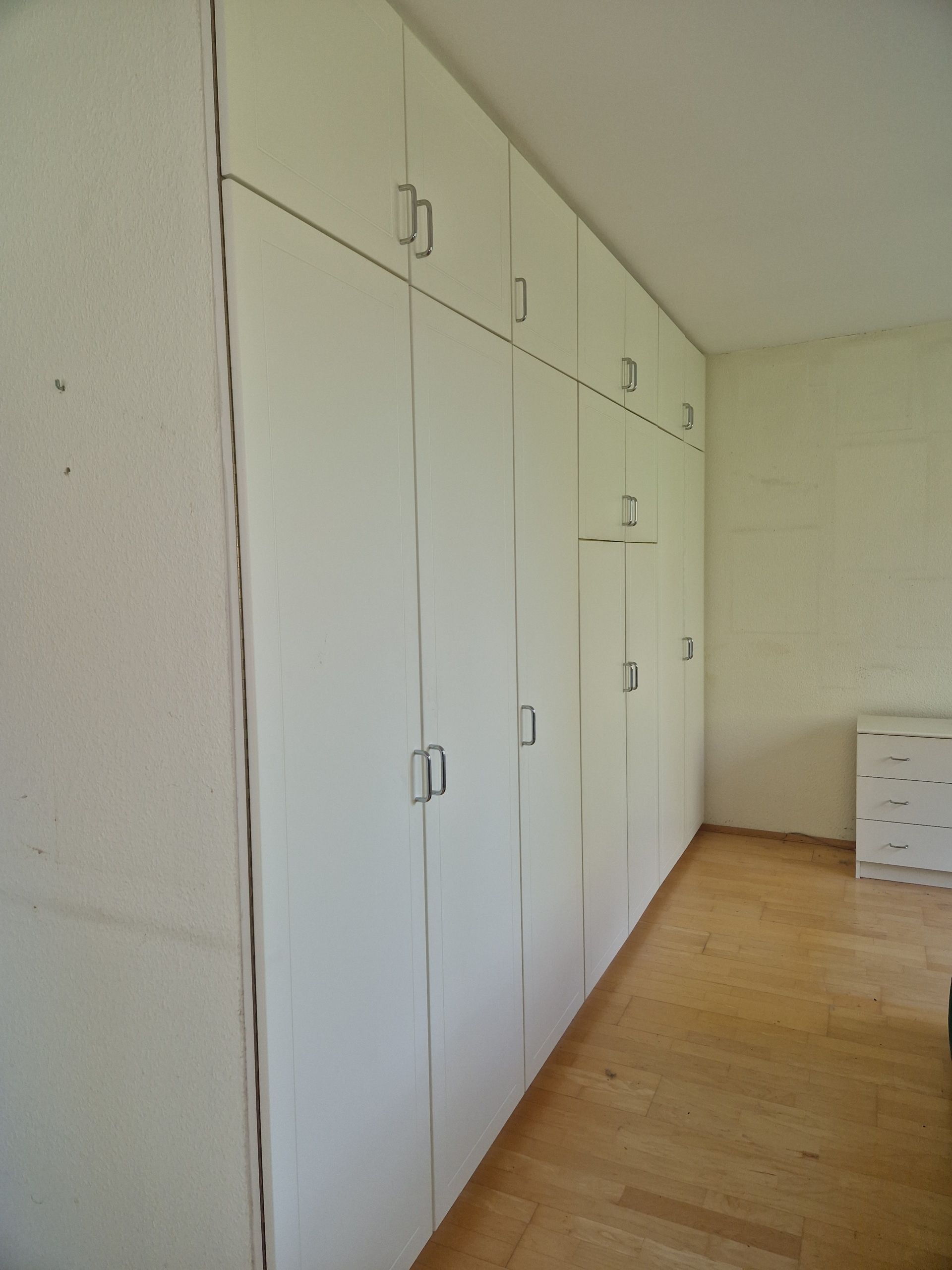 ein raumhoher weißer Einbauschrank mit silbernen Griffen unter schräger Decke auf Parkettboden