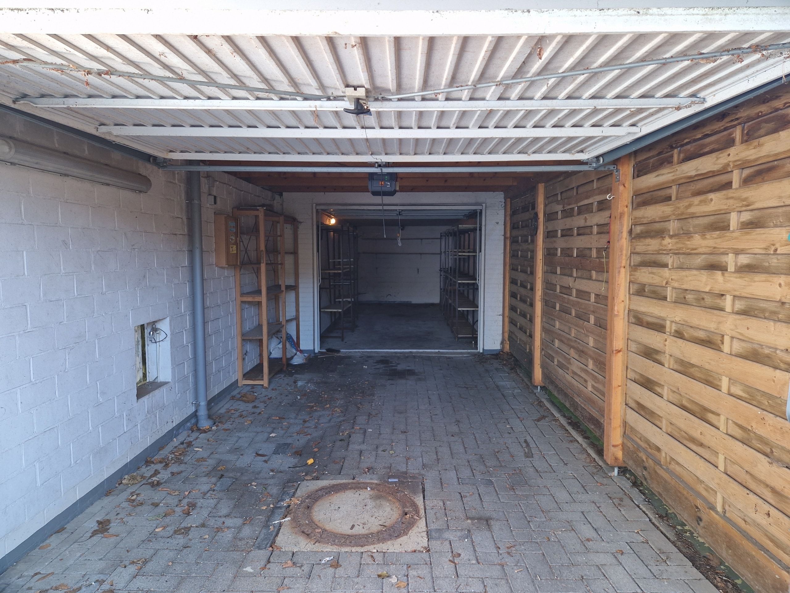 Offene Einzelgarage frontale Ansicht mit Pflasterboden links weiße Ziegelwand rechts Holzlattenverkleidung und Regalen