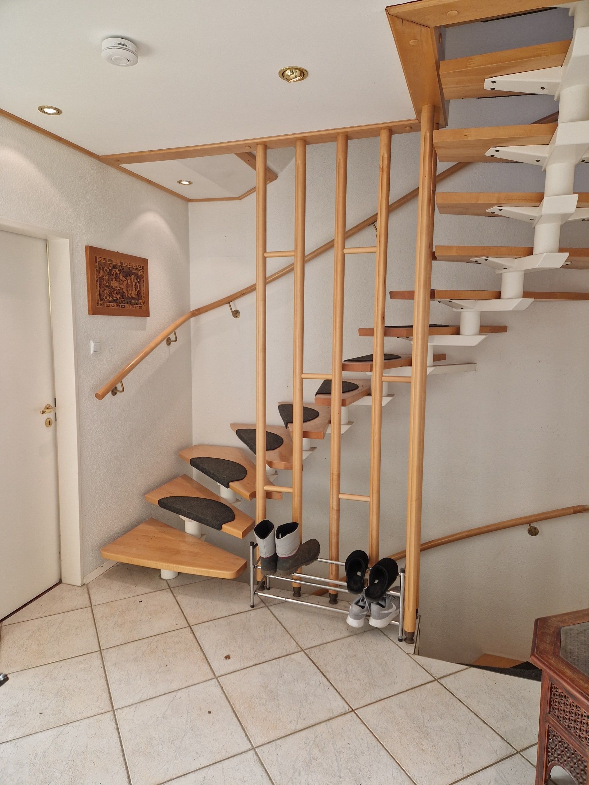 Eingangsbereich mit heller Buchenholz-Wendeltreppe an weißer Wand und grauem Fliesenboden mit Schuhregal