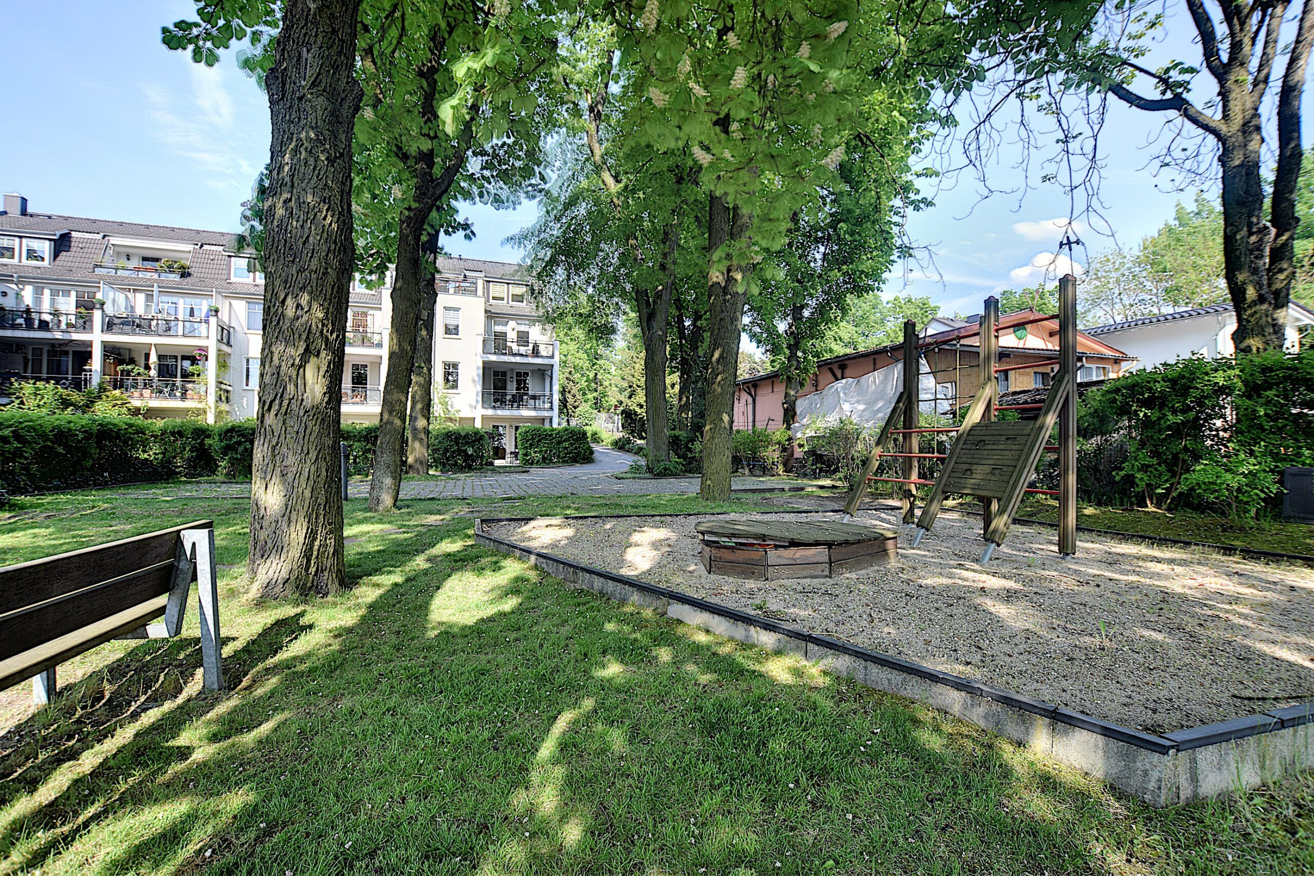 Mehrfamilienhaus mit weißen Balkonen und begrüntem Innenhof mit Sandkasten Klettergerüst und Parkbank aus frontaler Sicht