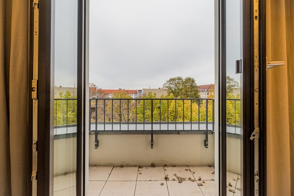 Widok wnętrza balkonu w mieszkaniu wielopiętrowym z podłogą wyłożoną płytkami, metalową balustradą i jesiennymi koronami drzew w tle