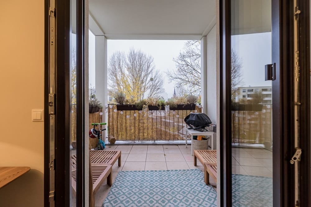 Widok przez otwarte drzwi salonu na zadaszony balkon z drewnianymi meblami ogrodowymi i porośniętą roślinnością balustradą